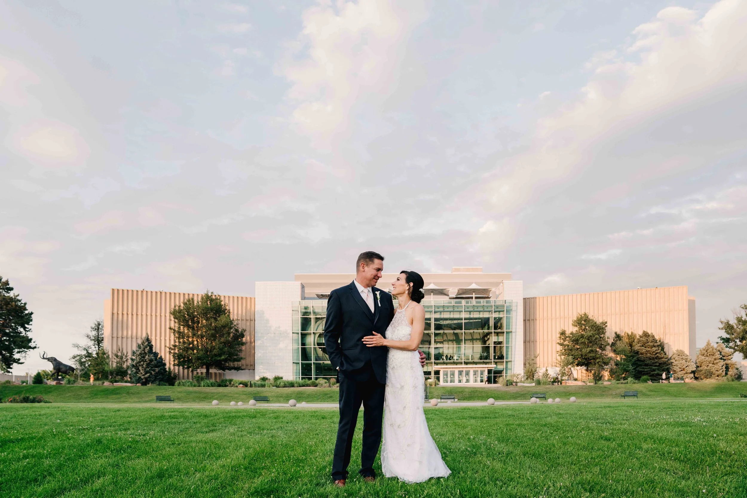 A bride and groom standing on a grassy lawn, gazing at each other, with a modern glass building and trees in the background, during sunset.