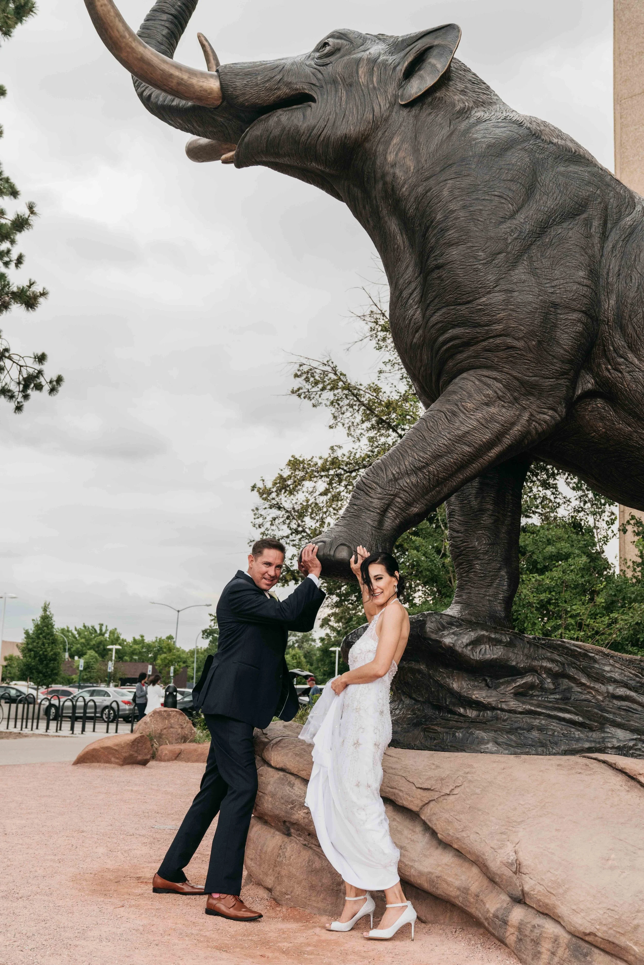 A couple in wedding attire standing under a large elephant statue, with the man holding the elephant's trunk and the woman smiling.