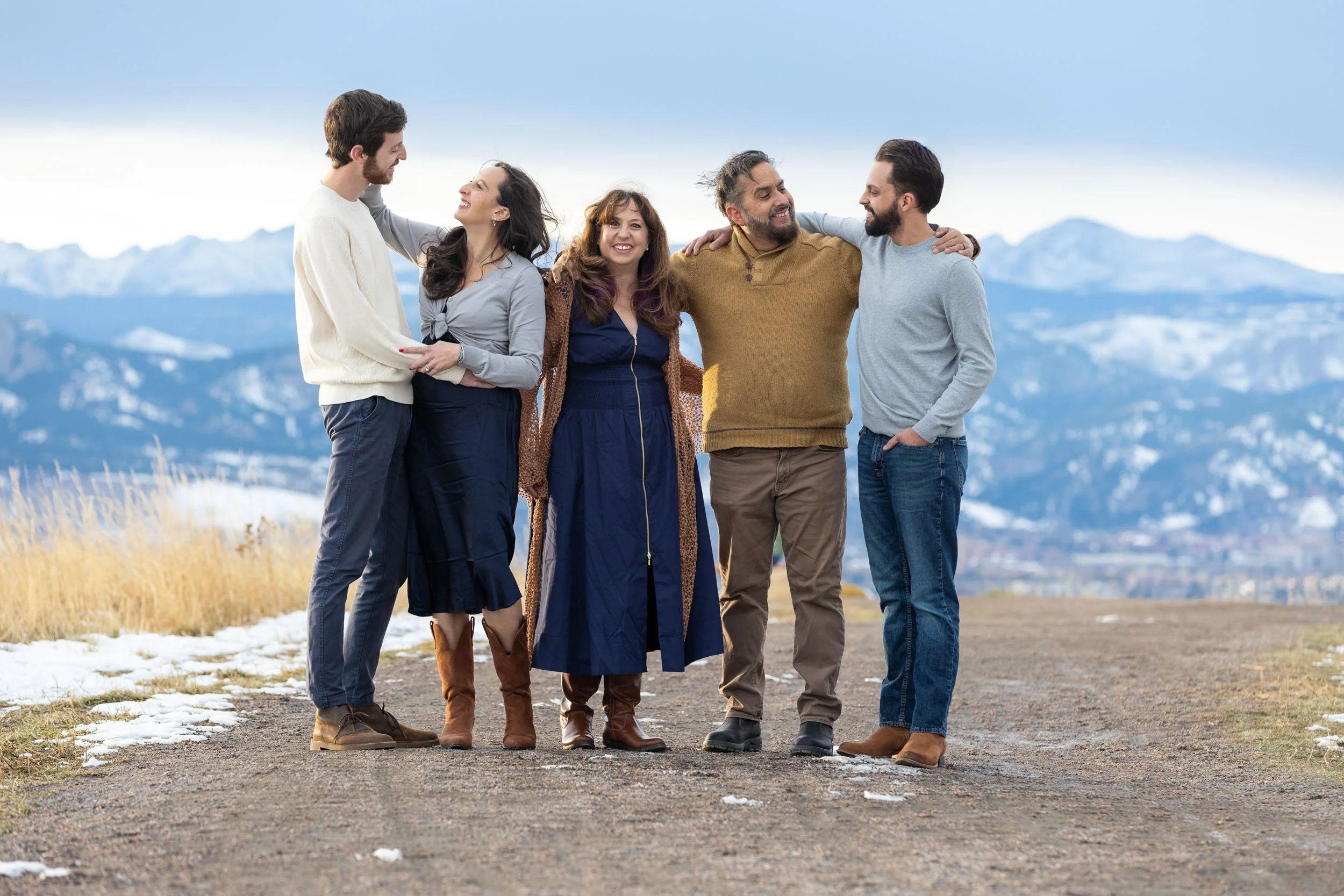 A group of six people standing on a dirt path outdoors, smiling and interacting, with snow-capped mountains in the background.