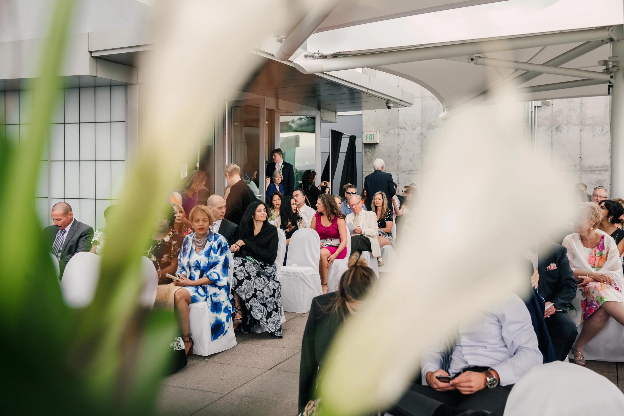 A diverse group of people seated on white chairs at an outdoor event, possibly a wedding or reception, with some people engaged with their phones or talking. The scene is partially obscured by blurred white flowers or plants in the foreground.