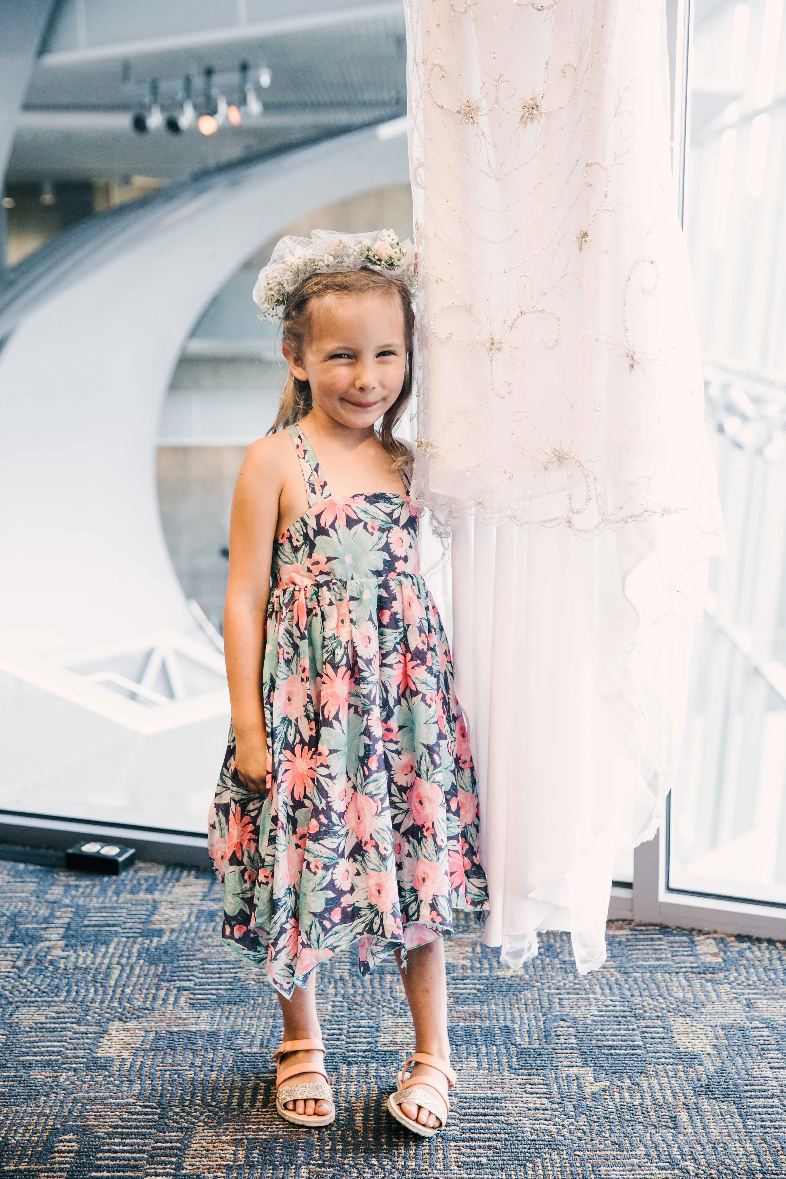 A young girl standing indoors near a white dress or curtain, wearing a floral dress, a flower crown, and sandals, smiling.