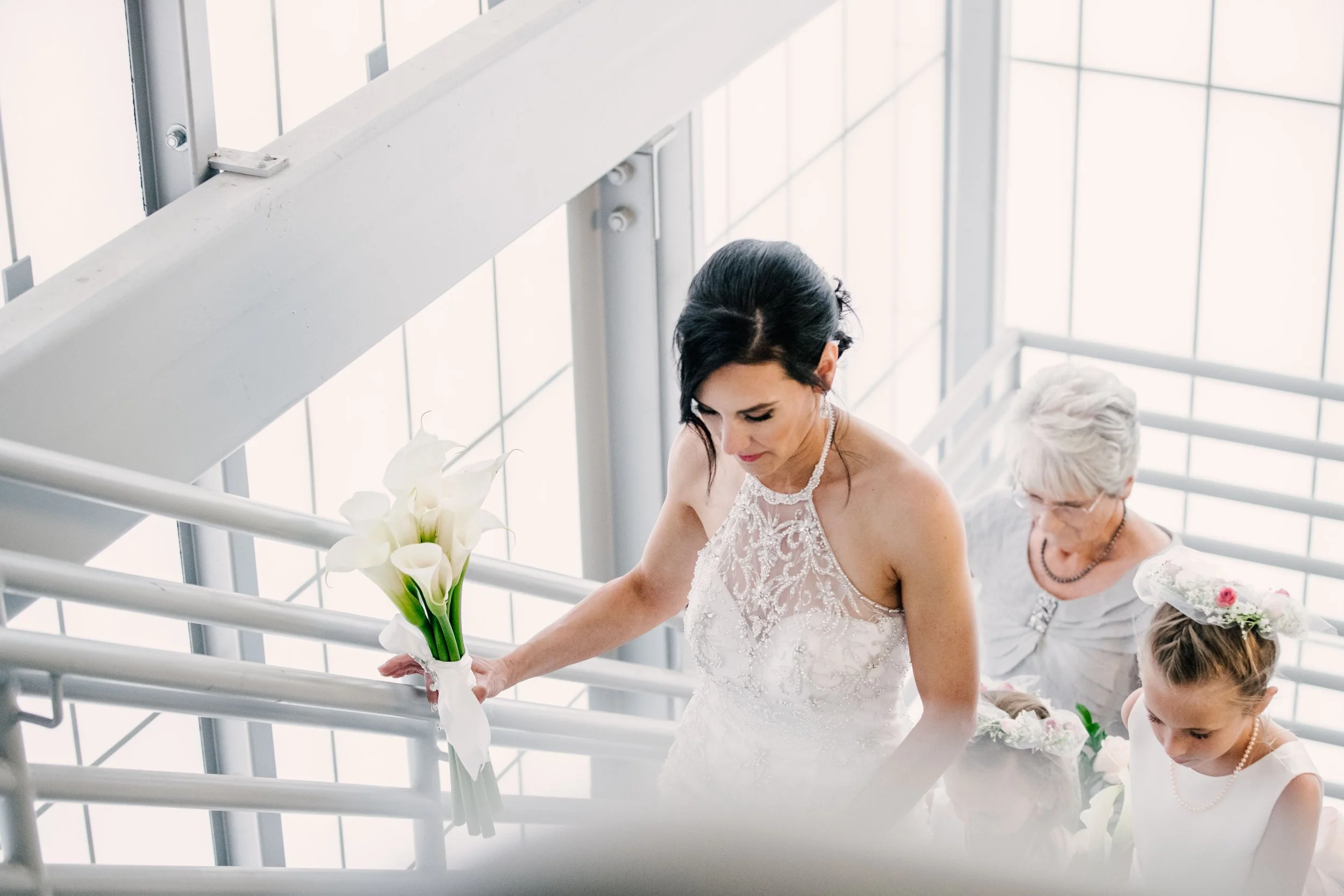 A bride with black hair in a white wedding dress holding a bouquet of white calla lilies climbing stairs with an older woman and a young girl, possibly a flower girl, following her up the staircase.