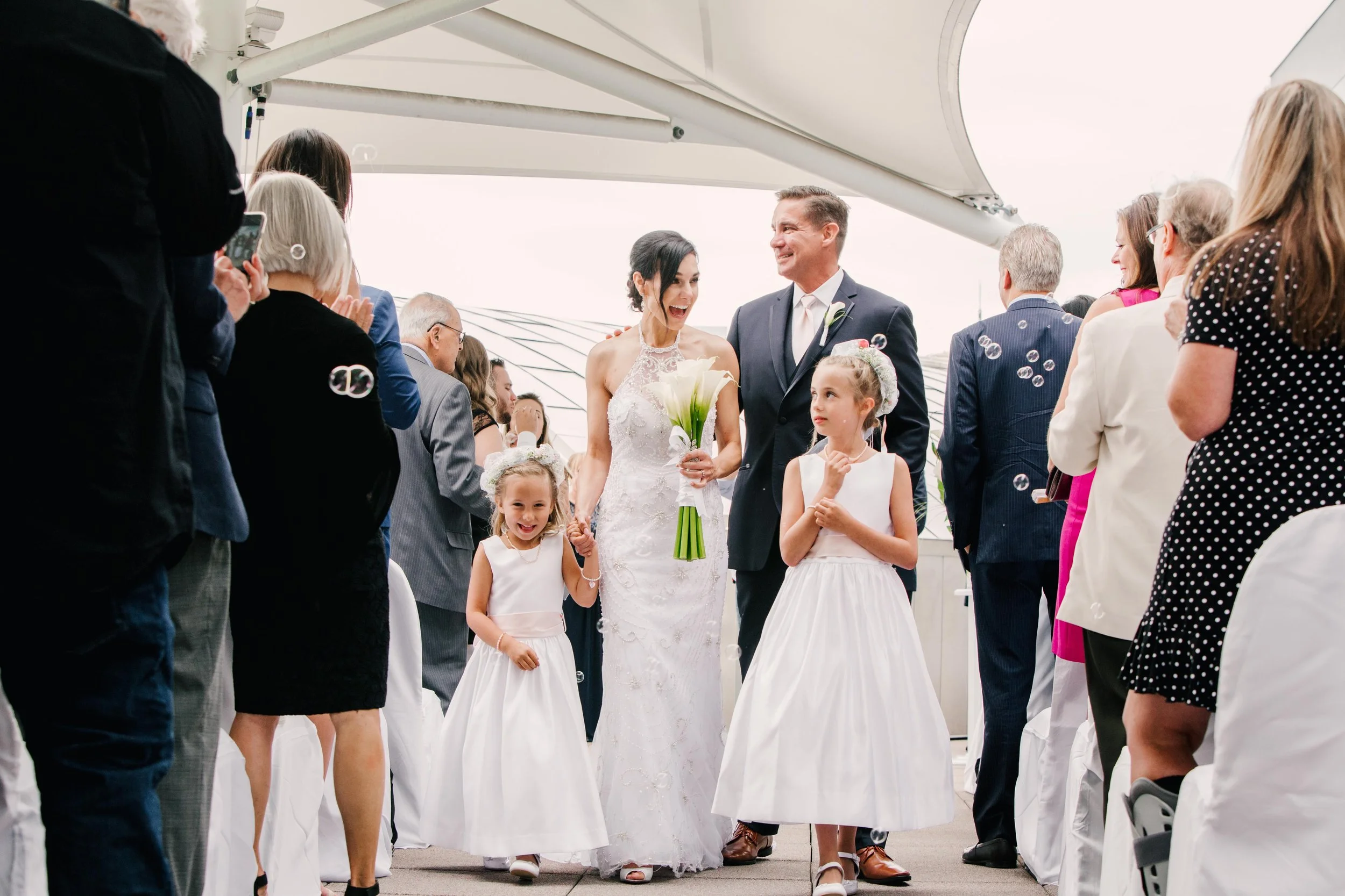 A bride and groom walking down the aisle at their wedding, surrounded by children and guests, with the bride holding a bouquet of white calla lilies, smiling and holding hands with one of the children, as bubbles are floating around.