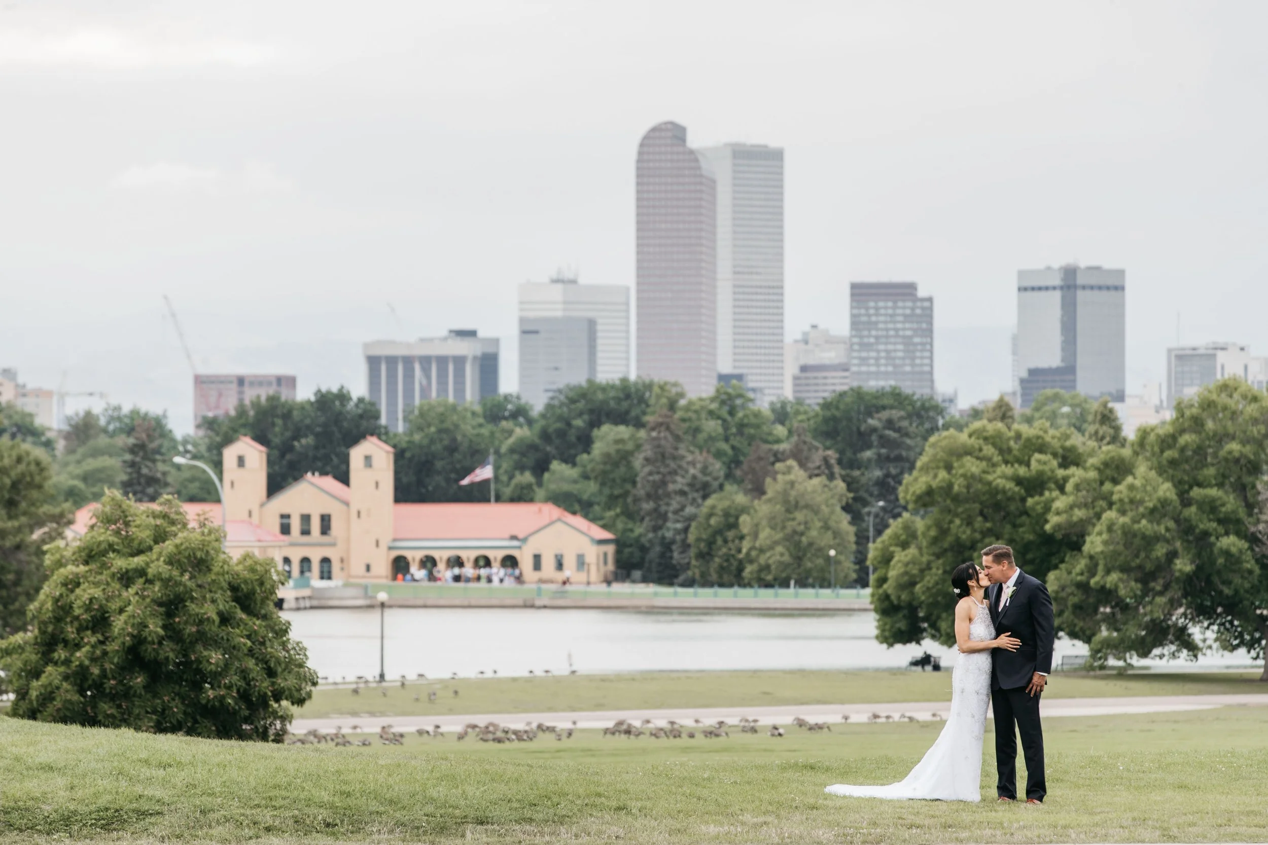 A bride and groom sharing a kiss in a park with city buildings in the background.