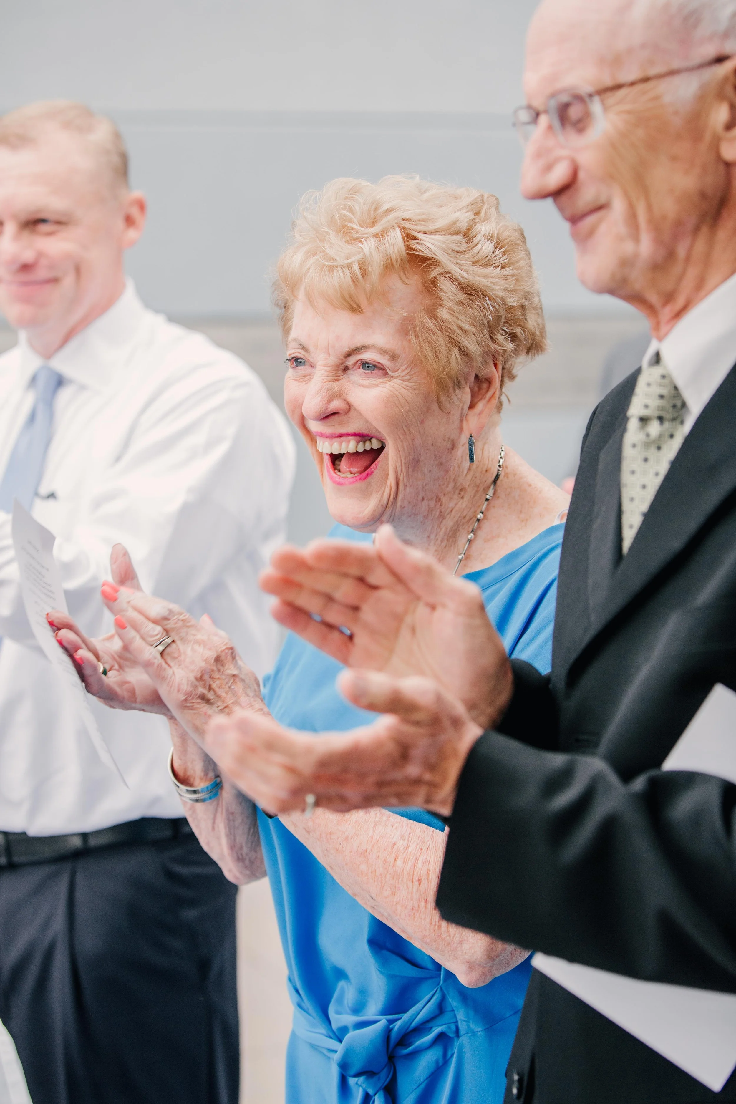 Three older adults, two men and one woman, are clapping and smiling, dressed in formal attire at what appears to be a professional or celebratory event.