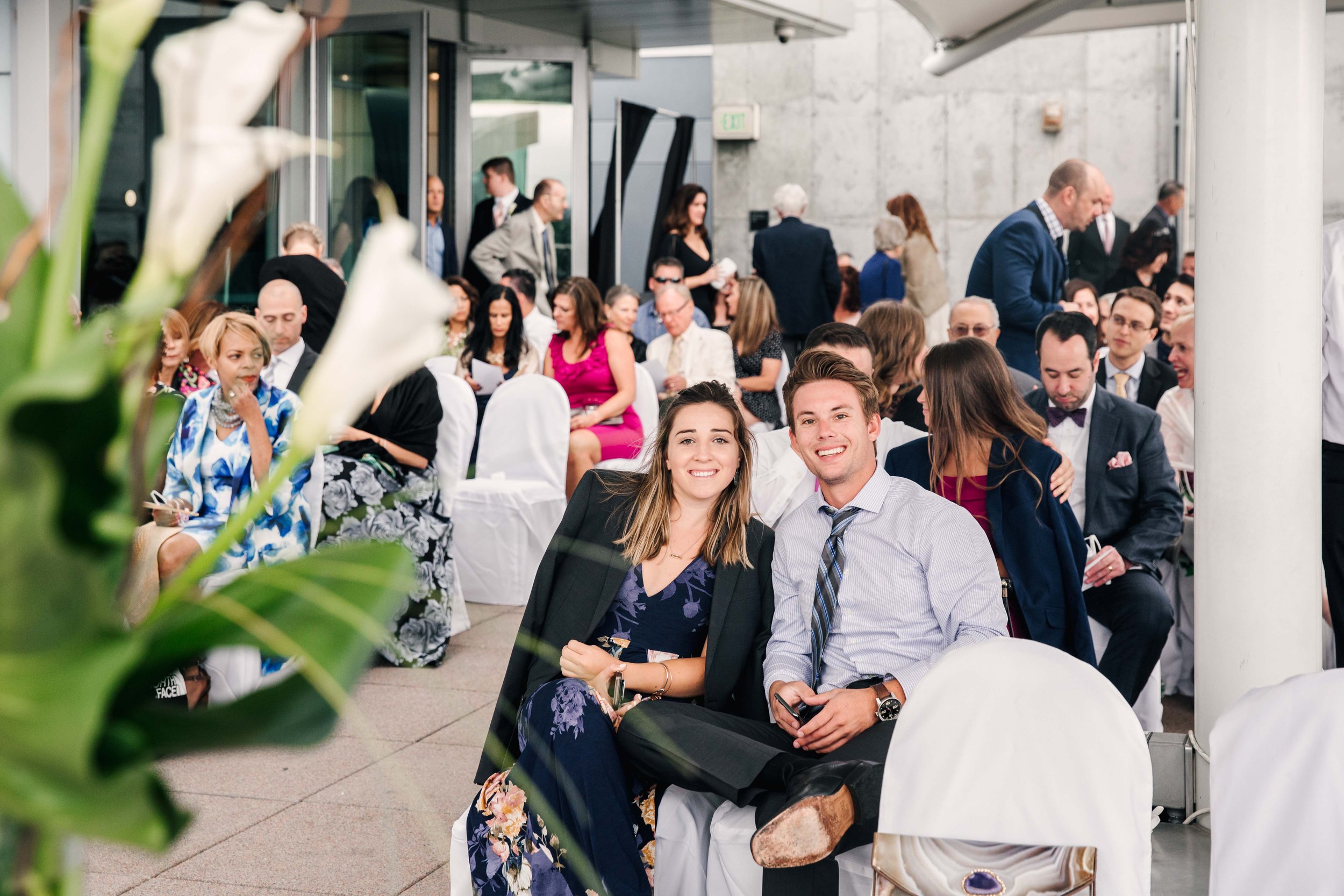 Two young adults, a woman and a man, sitting and smiling at a formal event with many people in the background.