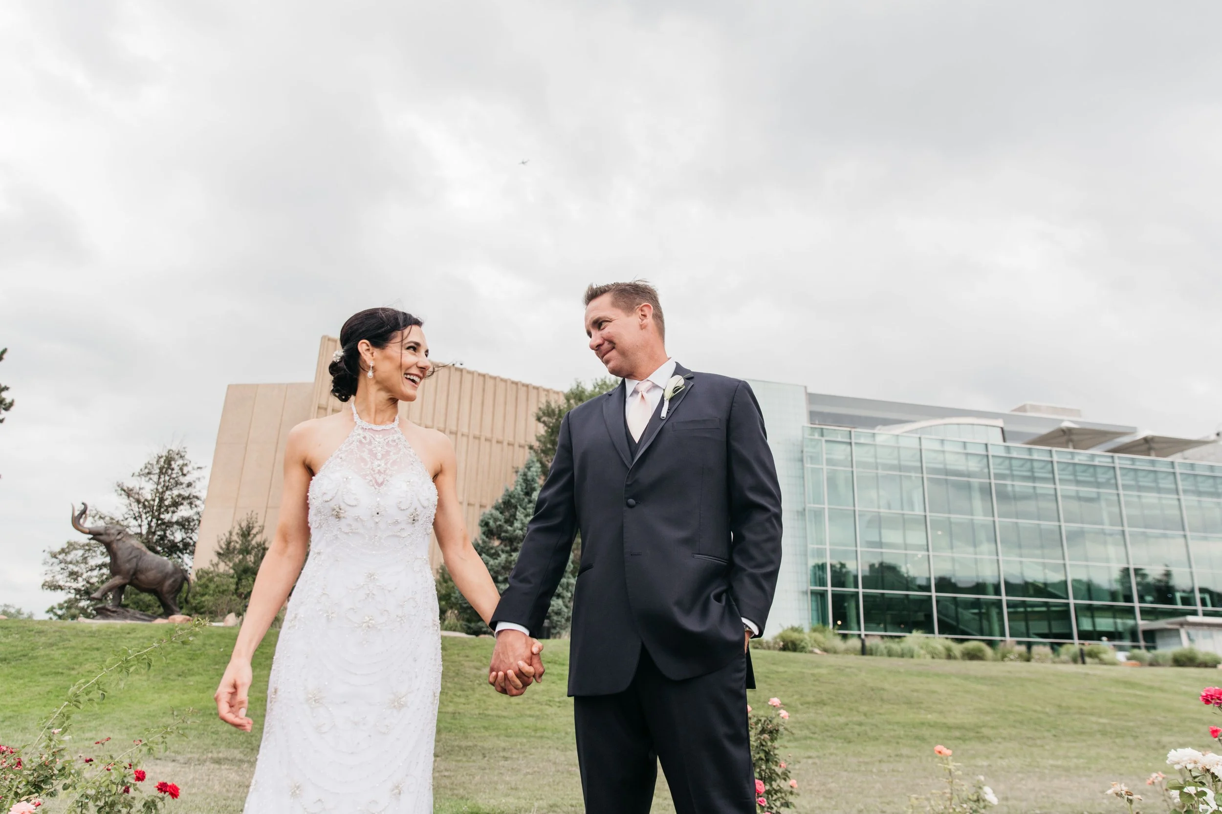 A bride and groom holding hands, smiling at each other outdoors in front of a modern building and cloudy sky.