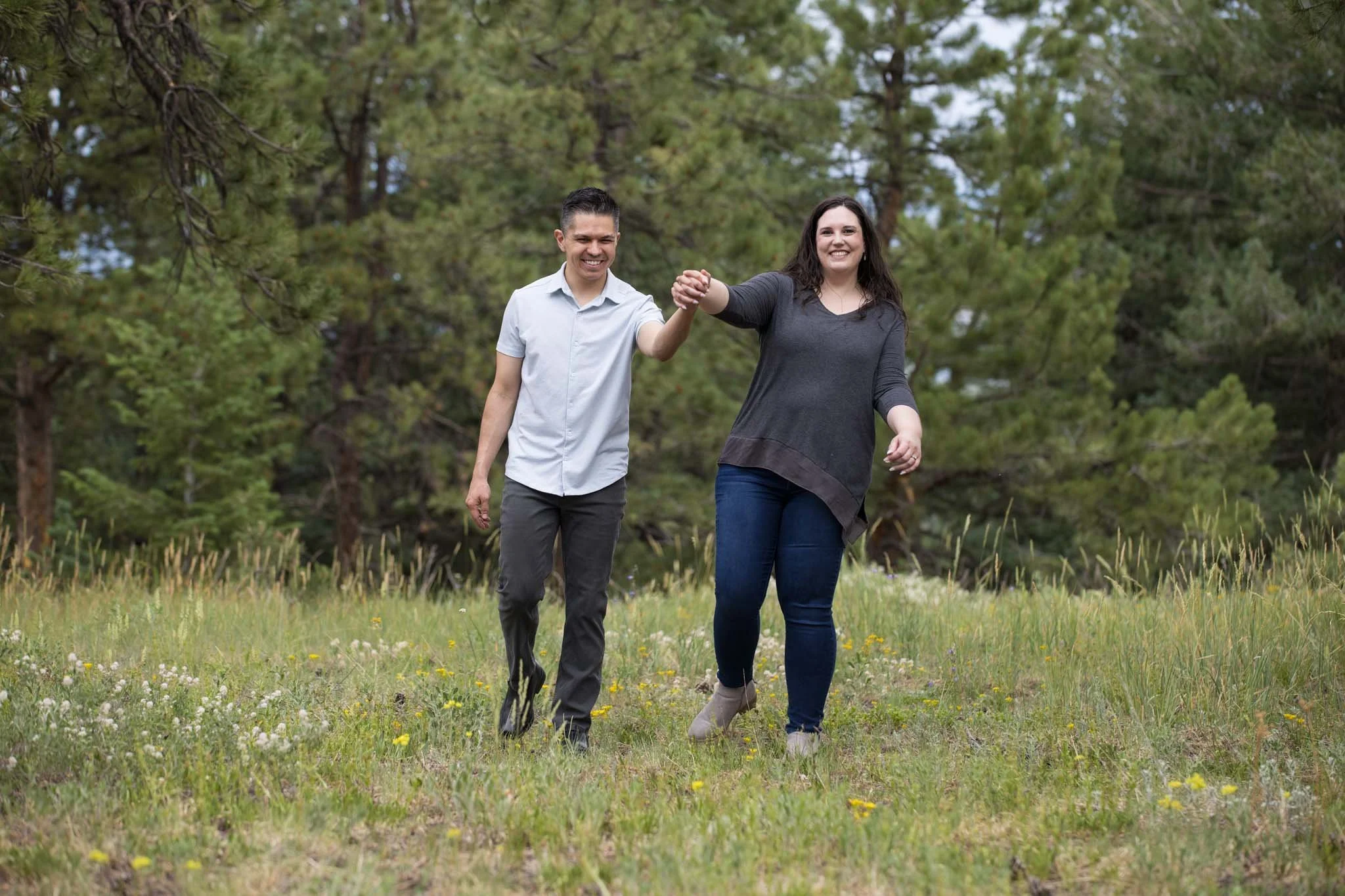 Couple holding hands while walking on grassy field with trees in background.