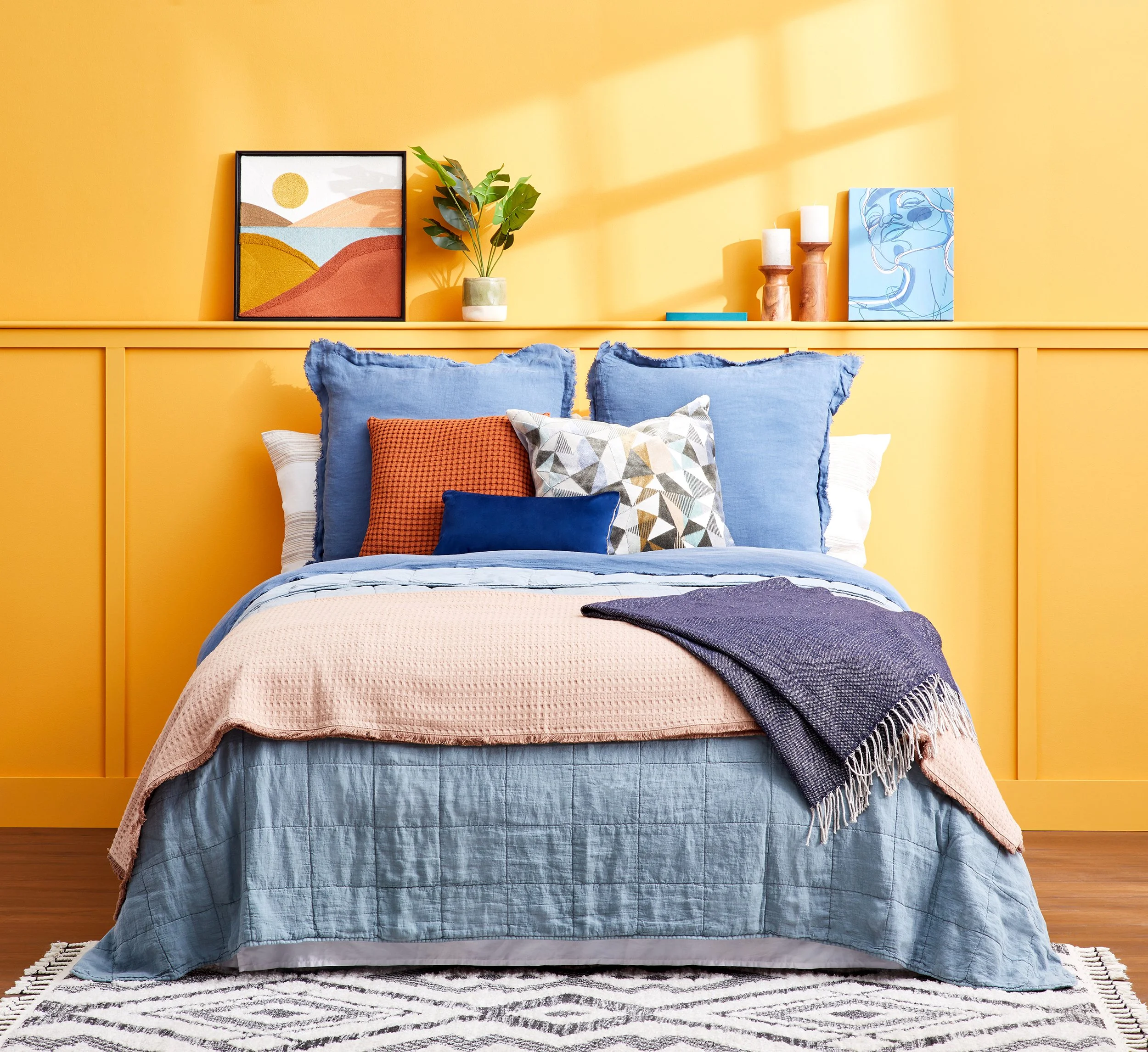 Adam Krauth Photo - Commercial Photography - Boston, MA - Still life - Interior photography. A scene with a bed against a bright yellow wall. Decor includes artwork, a potted plant, candles, and a striped rug.