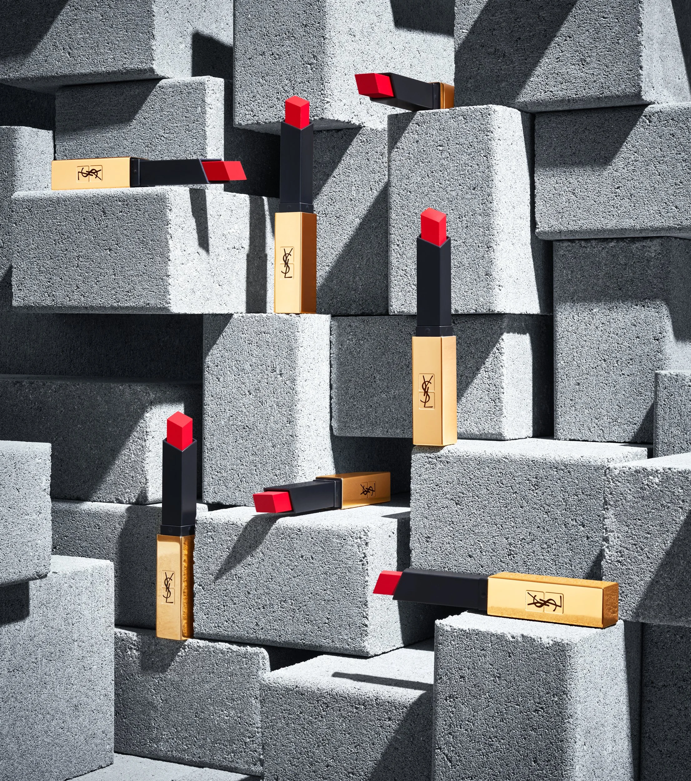 A set of abstract designer lipstick tubes, painted in black and gold with red caps, arranged on a textured concrete wall with shadows cast across. The tubes have a stylized logo, reminiscent of Yves Saint Laurent.