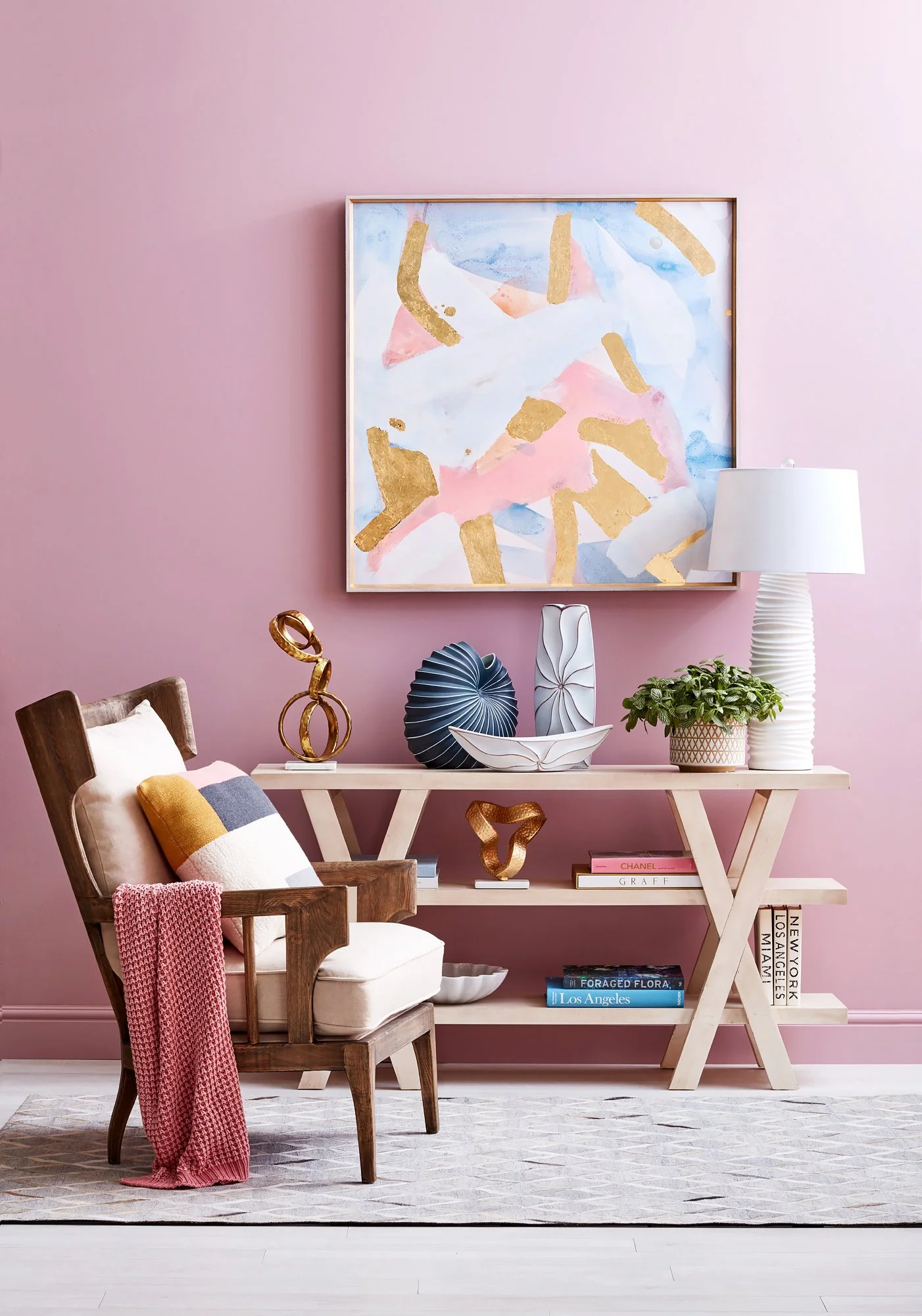 Adam Krauth Photo - Commercial Photography - Boston, MA - Still life - Interior photography. A scene with an interior of a room with a pink wall, featuring a wooden armchair and shelving with pastel colors and gold accents hanging on the wall.