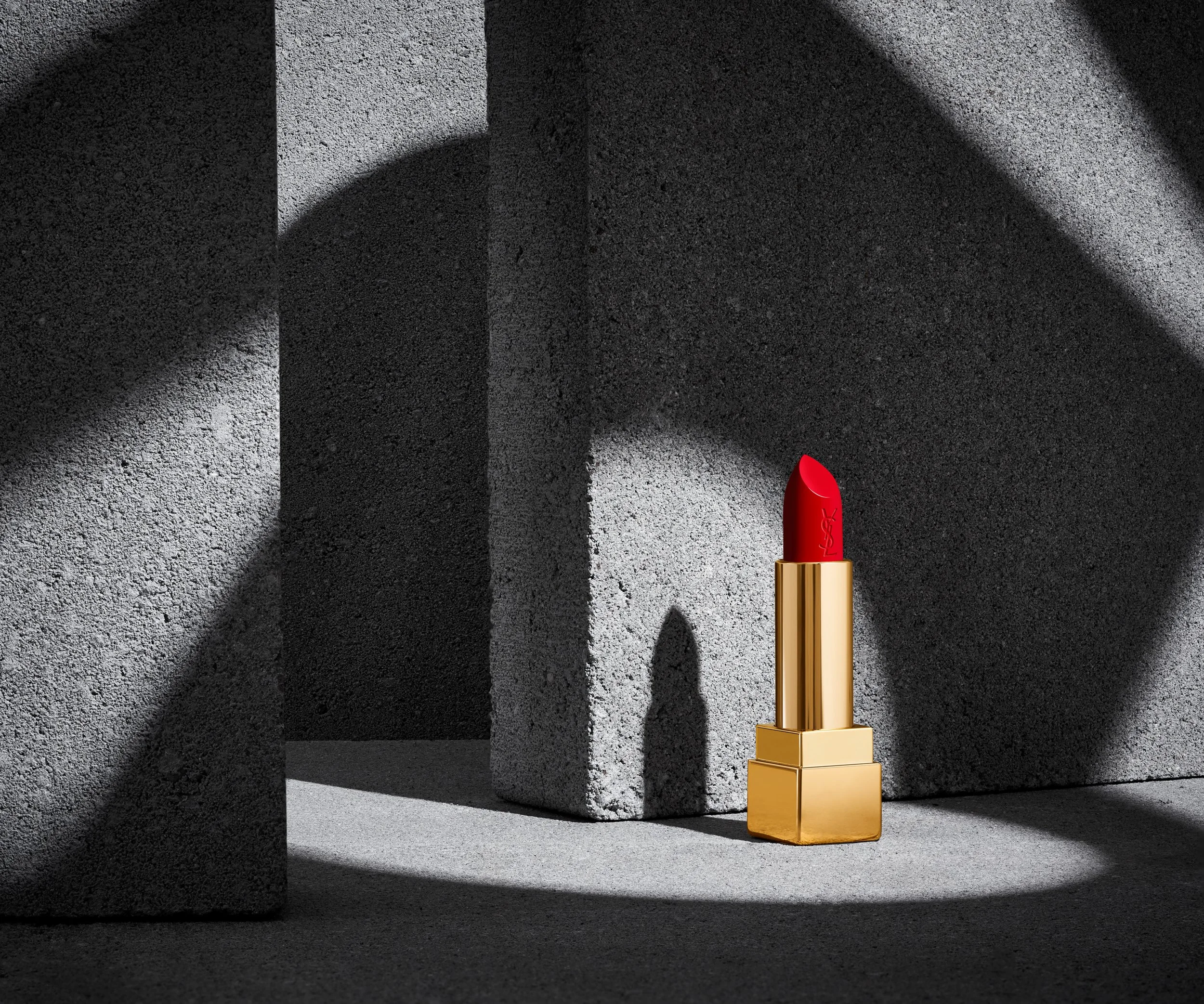 Adam Krauth Photo - Commercial Photography - Boston, MA - Still Life - Product Photography - A red lipstick tube with a golden base standing on a gold platform, casting shadows on a textured gray background. Cosmetics photography 