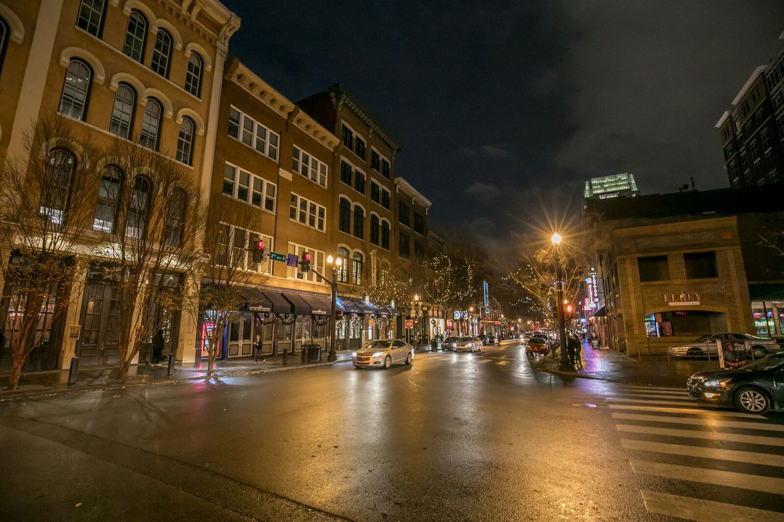 Nighttime view of a city street with historic buildings, lit by streetlights, decorated with string lights, and moving cars on a wet road.