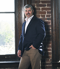 Man in a navy blazer and beige pants standing by a large window with a brick wall background.