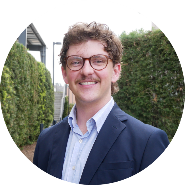 Sean Iverson, a young man with curly brown hair, wearing glasses, a light blue button-up shirt, and a dark blazer, standing outdoors with greenery and stairs in the background, smiling at the camera.