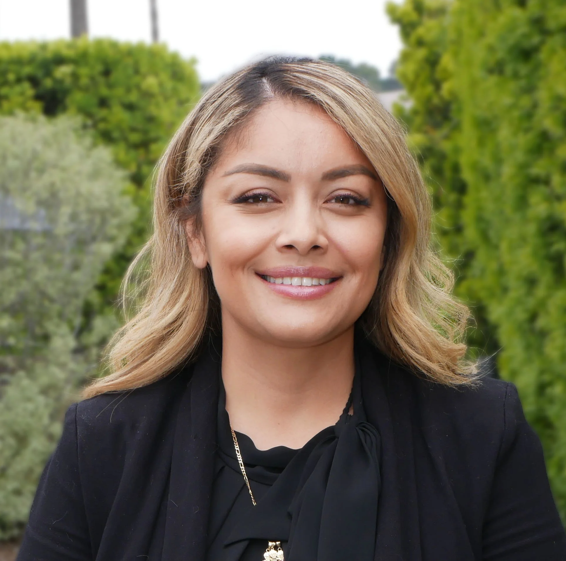 A woman smiling outdoors with green foliage and trees in the background.