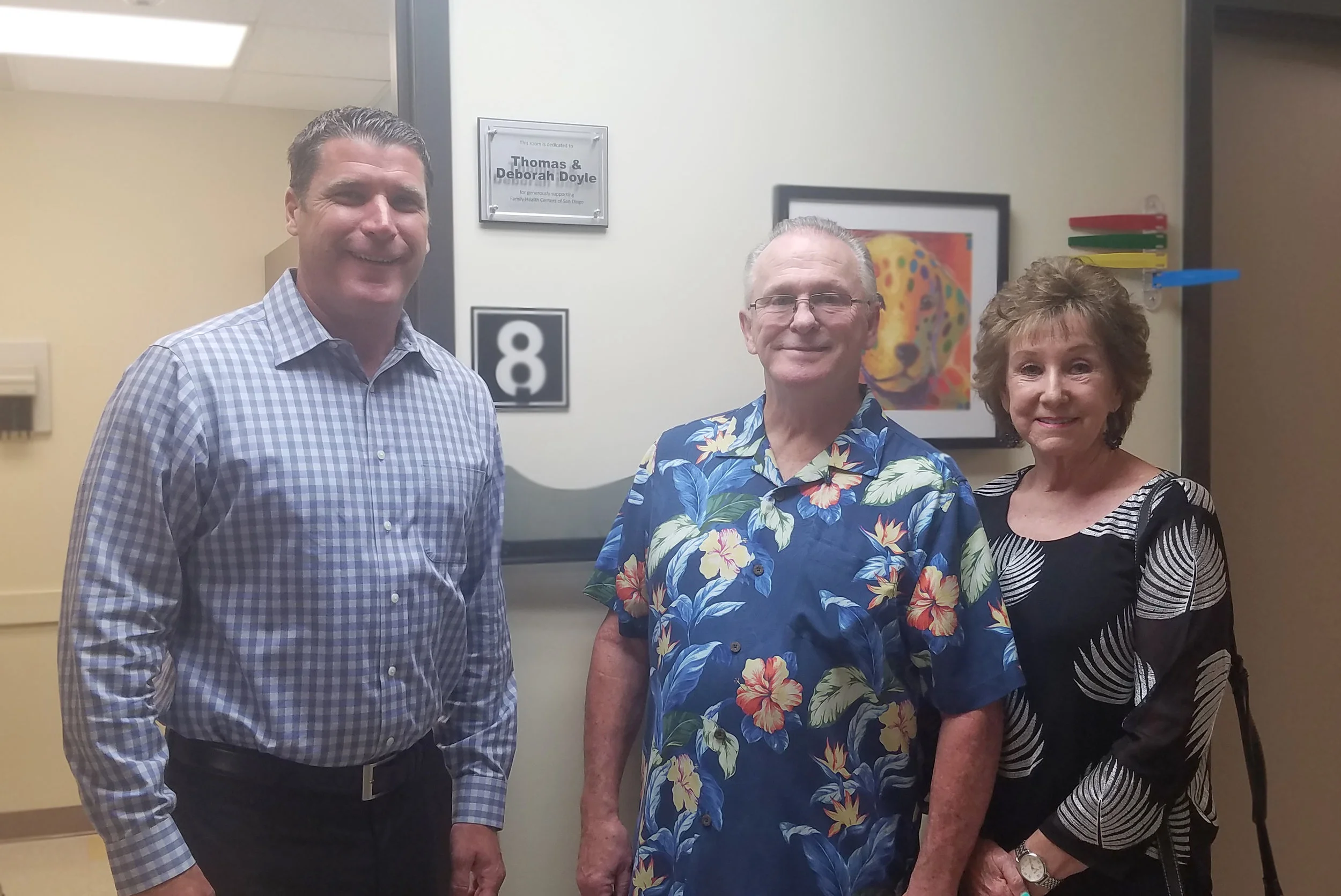 Sam Clayton, Tom & Debby Doyle standing in front of the room plaque.