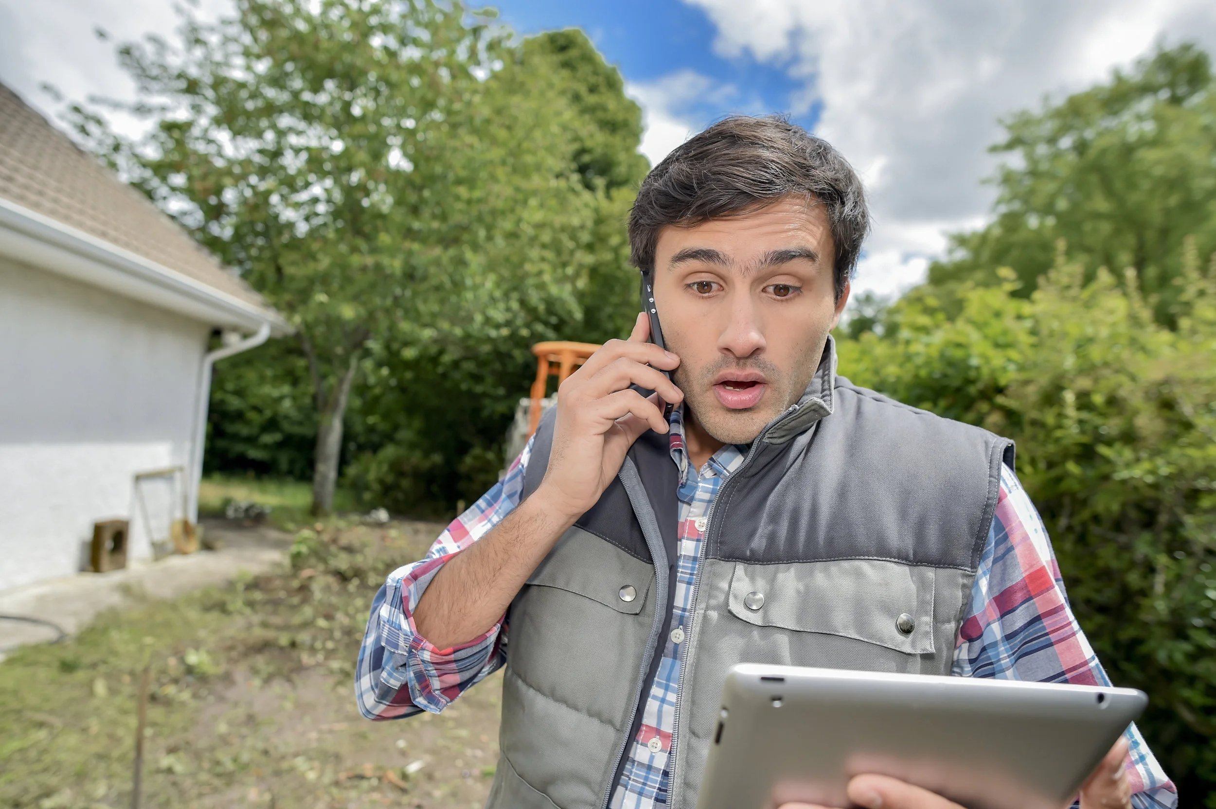 Man talking on mobile phone while looking at a tablet computer standing outside among landscaping.