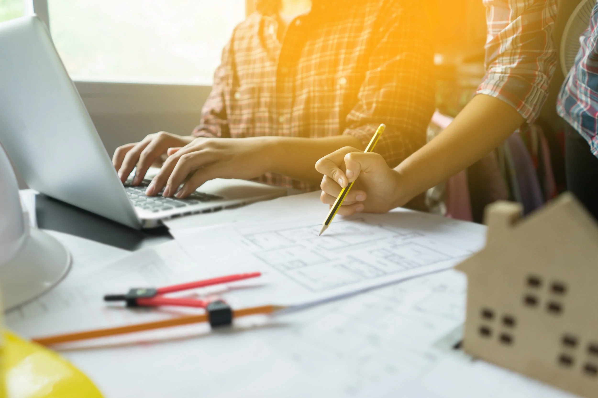 Man at a desk working on a laptop with a women standing next to him holding a pencil to a floorplan on the desk.
