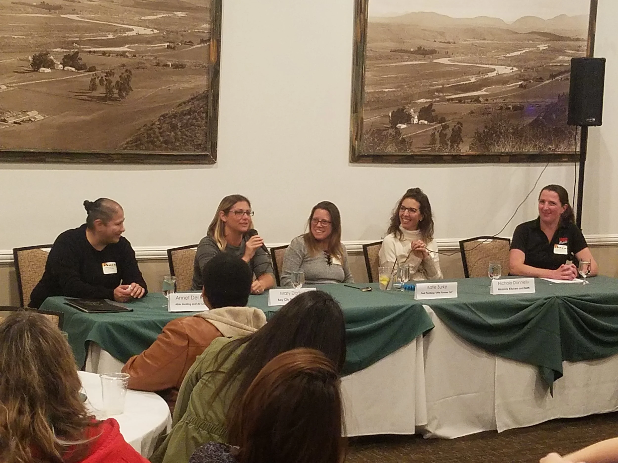 Five women sitting at a rectangular table with one holding a microphone