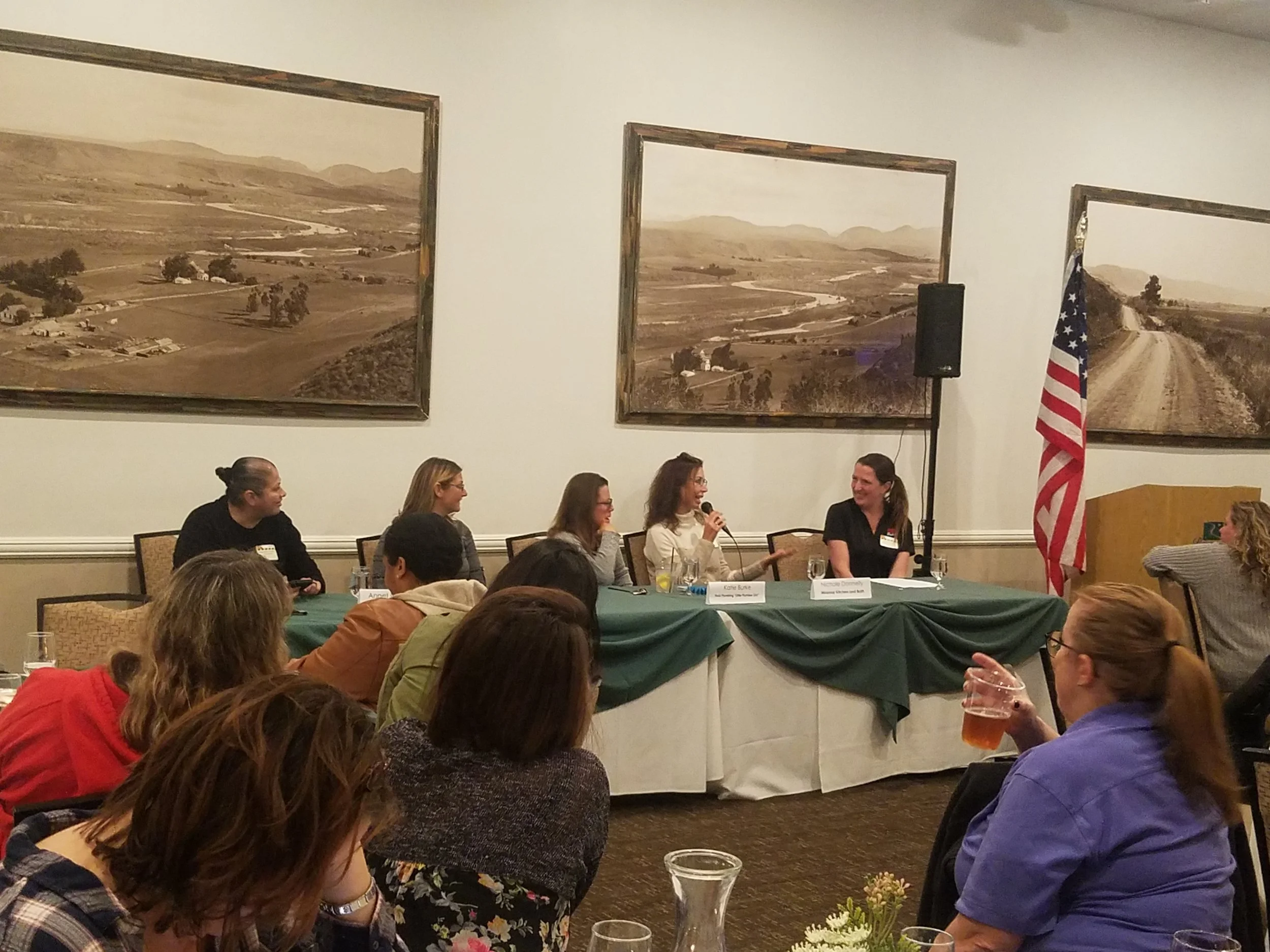 Five women sitting at a rectangular table with one holding a microphone