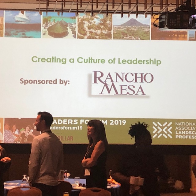 Drew Garcia and Margaret Hartmann standing at their table with the giant screen with Rancho Mesa logo in the background