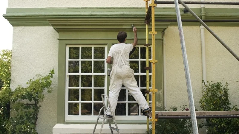 Guy standing one leg on ladder and one leg on scaffold while painting house.
