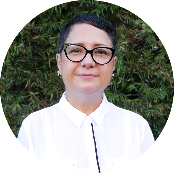 Elia Gonzalez, a woman with short dark hair, wearing glasses, a white collared shirt, earrings, and standing in front of green foliage.