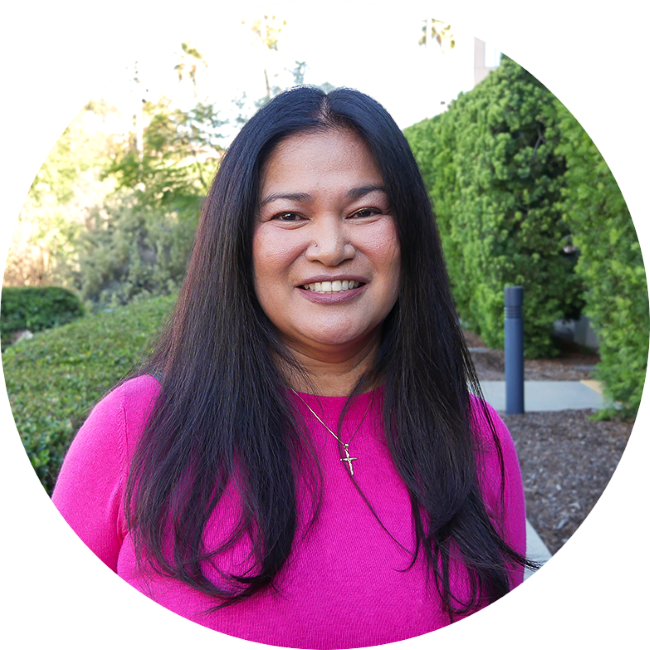Joyce Flores, a woman with long black hair smiling outdoors, wearing a pink top and a necklace with a cross pendant, in front of green bushes and trees.