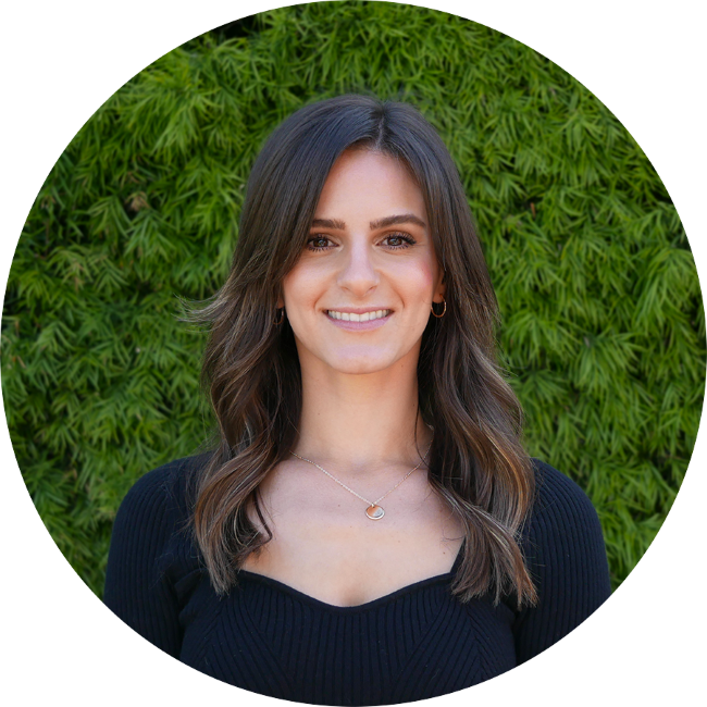 Sierra Stone, a woman with long, wavy brown hair smiling in front of green shrubbery, wearing a black top and silver jewelry.