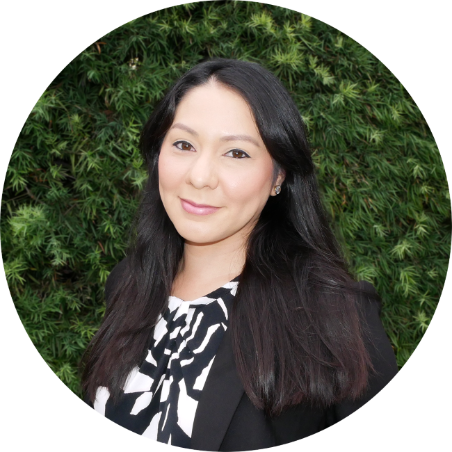 Georgina, Diaz, a woman with long black hair smiling in front of green foliage background, wearing a black blazer and a patterned top.