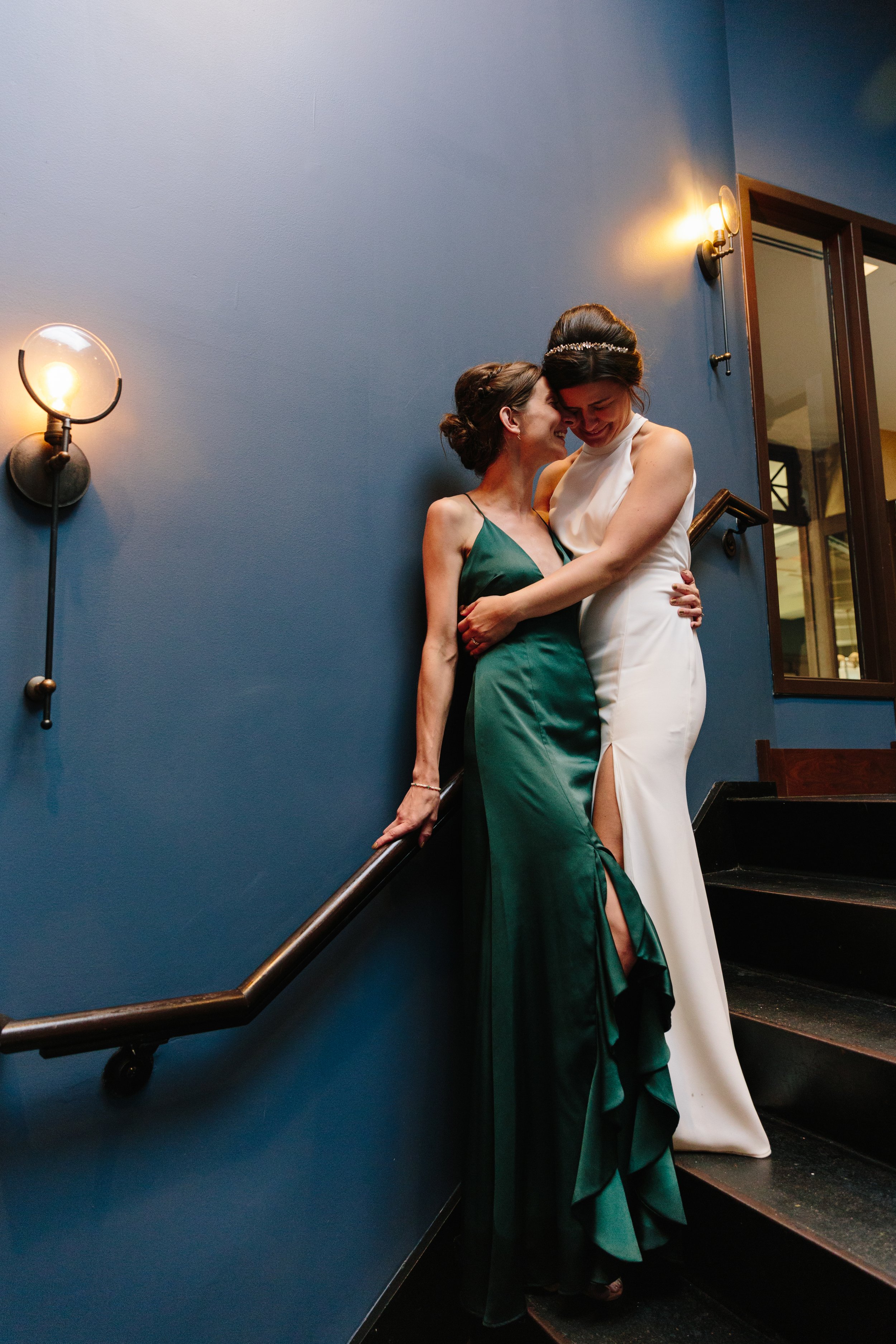  Candid documentary wedding photo of a bride in long white dress and bridesmaid in long stain green dress, laugh and hug in stairwell of intimate wedding reception. They stand in front of a blue wall and lights at Firehouse Restaurant in Chicago’s So