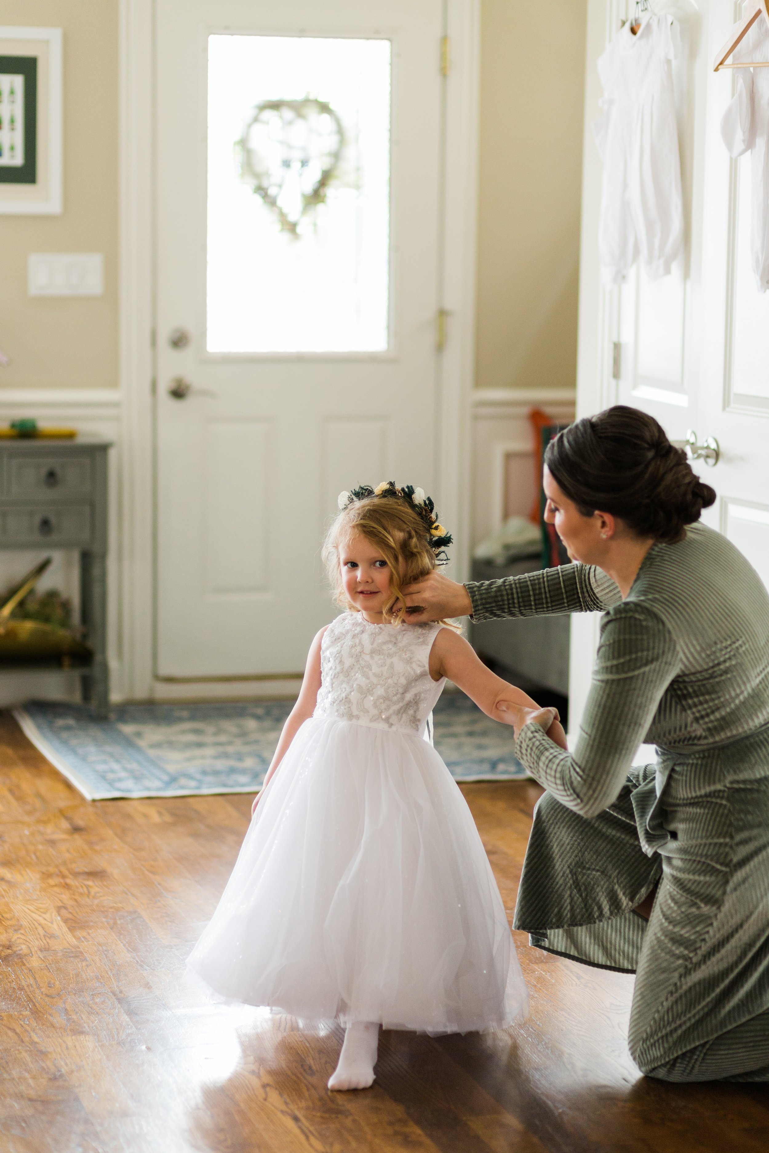  Candid documentary style wedding photo of a bridesmaid in a green velvet dress helping a young flower girl into her white dress while getting ready for an intimate wedding at home in Portage Park Chicago. 