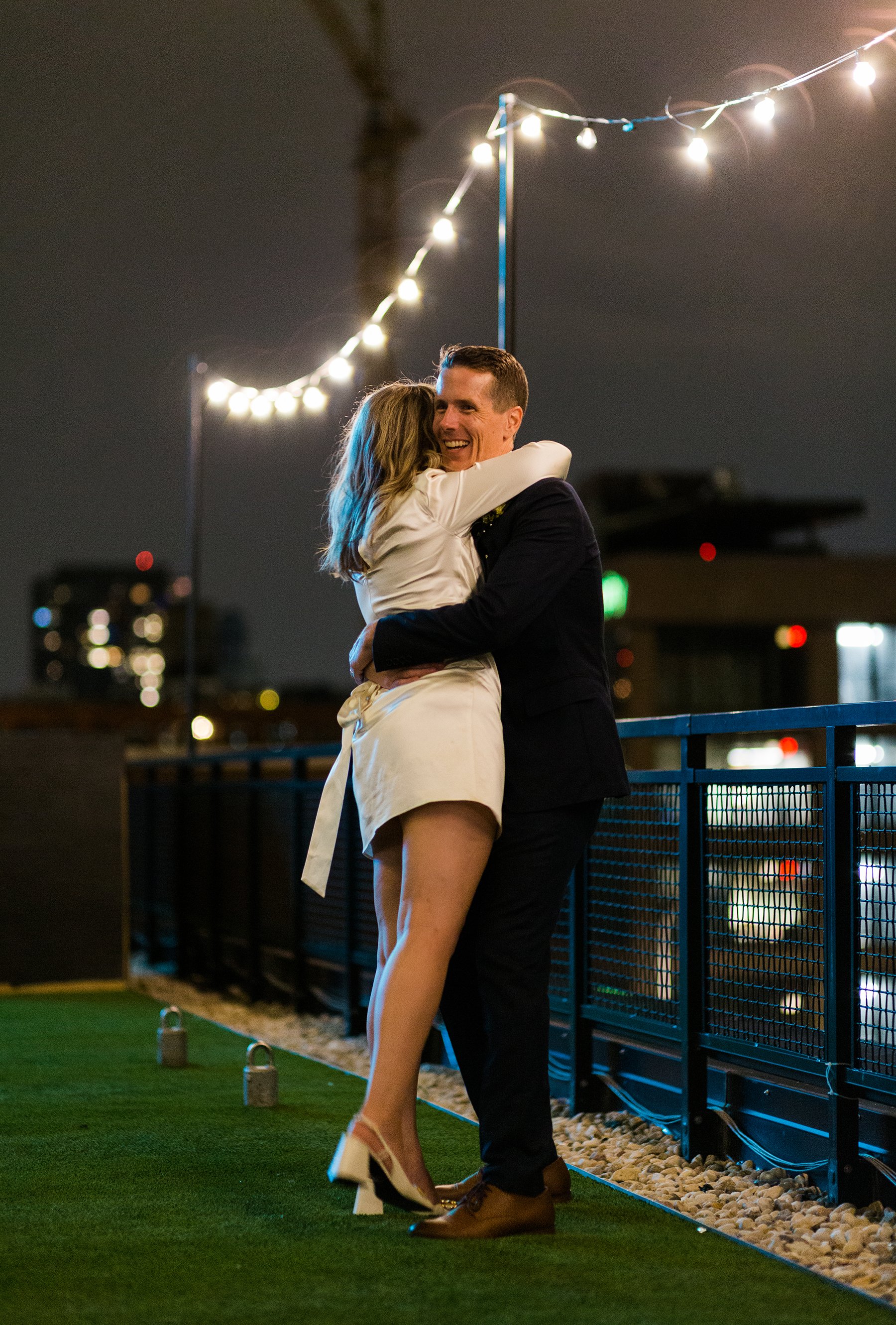  Candid documentary style photo of nontraditional bride in a short white dress being embraced by a smiling groom at the intimate night time wedding reception on the roof of The Emily Hotel in West Loop Chicago.  