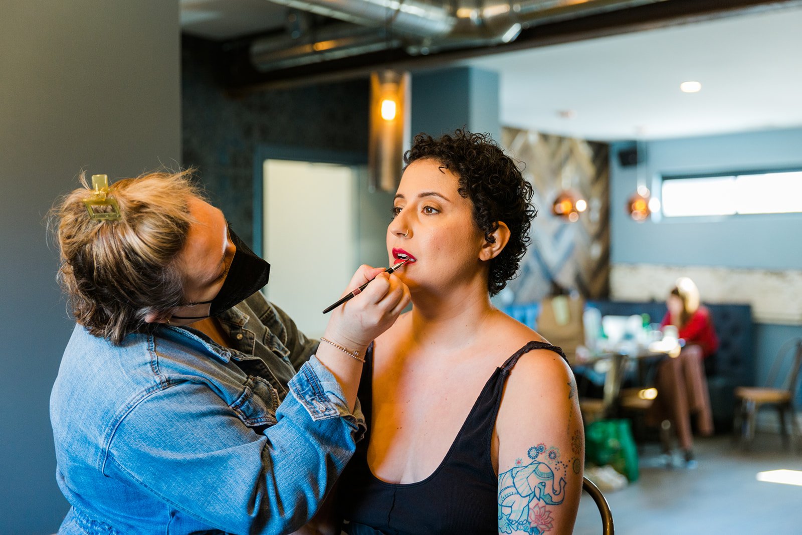  Documentary style photo of Nontraditional, tattooed, queer bride with short curly hair gets make up done by make up artist khaki pixley before wedding  