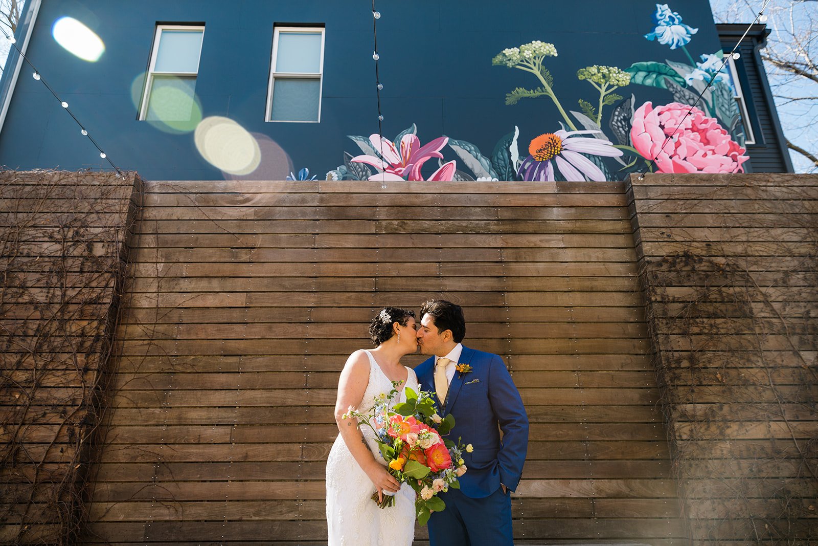 Nontraditional bride and groom kiss in front of a floral mural with a rainbow sun flare above them. 