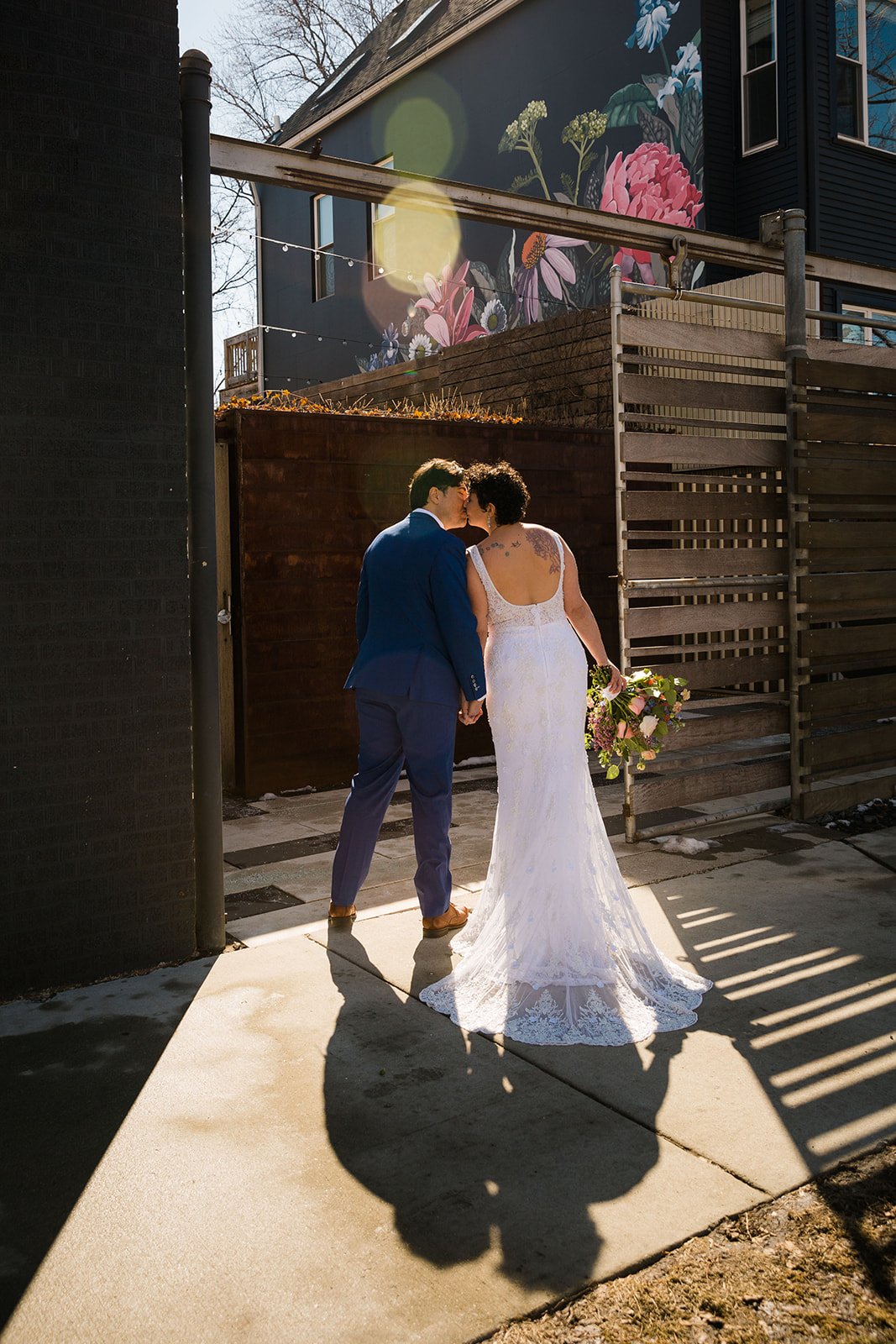  Portrait of nontraditional wedding from behind kissing in the patio doorway at The Joinery Chicago in Logan Square. Their shadow is behind them on the sidewalk and a colorful sun flare emerges from the top. Bright, colorful, unique wedding photograp