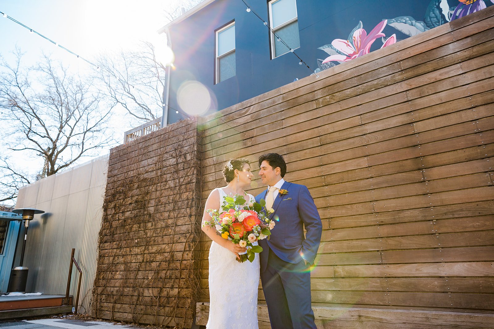  Portrait of nontraditional wedding couple looking at one another. Queer bride with short curly hair is in a white dress with a large colorful wedding bouquet and latino groom is in a blue suit with a yellow tie. They stand in front of a wooden panel