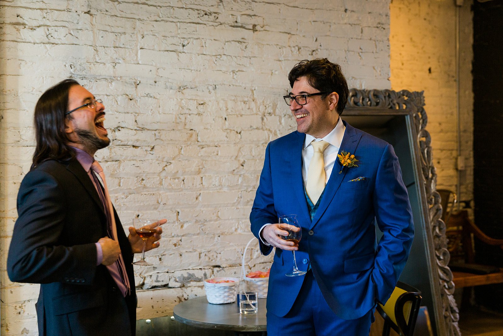  Documentary candid photo two latino men who are best friends laughing  with cocktails before nontraditional queer, Jewish and Venezuelan wedding ceremony at The Joinery Chicago in Logan Square. 