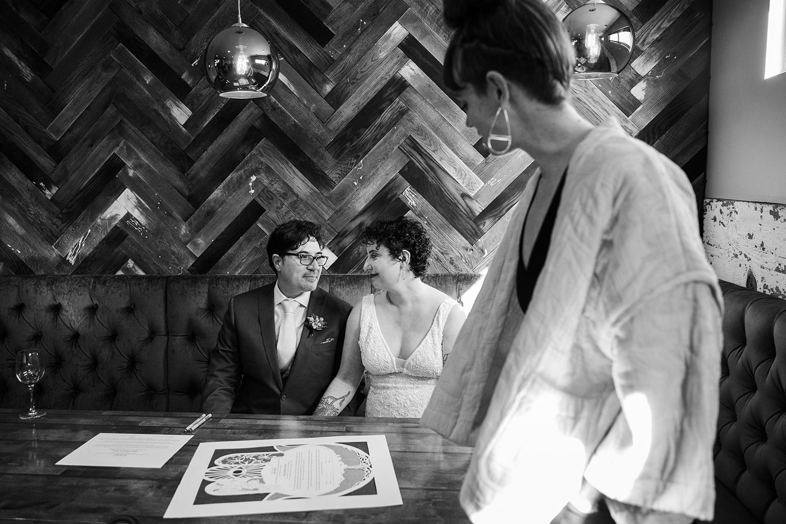  Black and white Documentary candid photo of the bride and groom looking at one another during their ketubah ceremony before nontraditional queer, Jewish and Venezuelan wedding ceremony at The Joinery Chicago in Logan Square. 