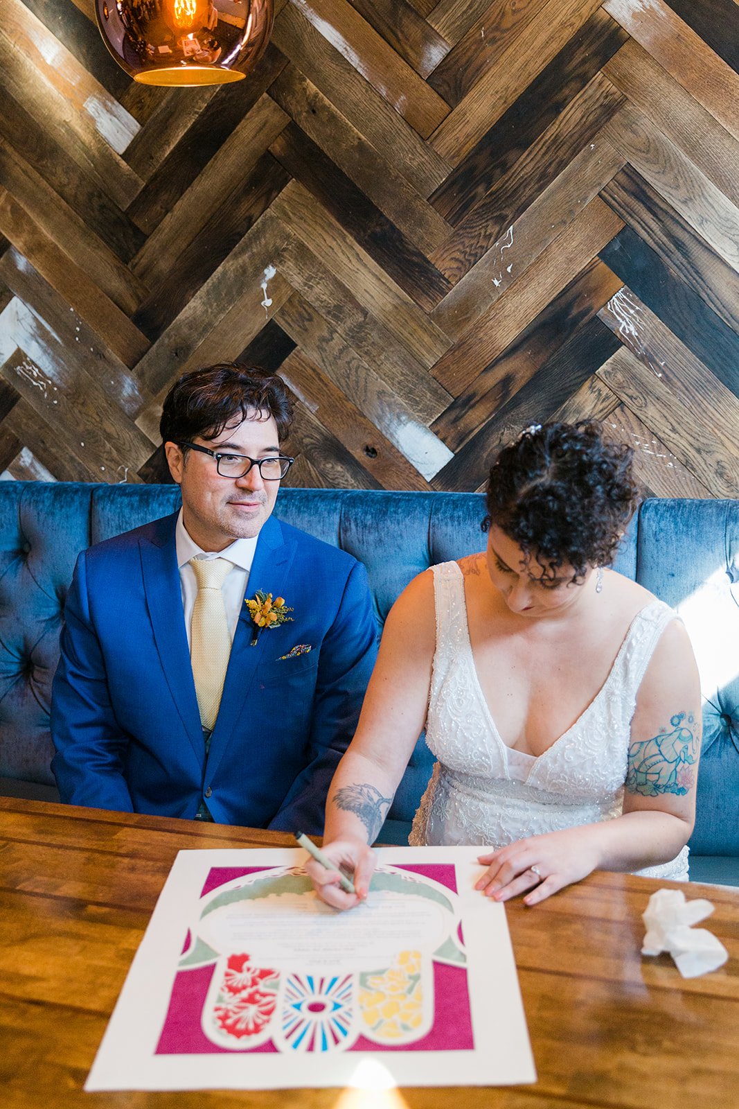  Documentary candid photo of the bride and groom signing their ketubah signing before nontraditional queer, Jewish and Venezuelan wedding ceremony at The Joinery Chicago in Logan Square. 