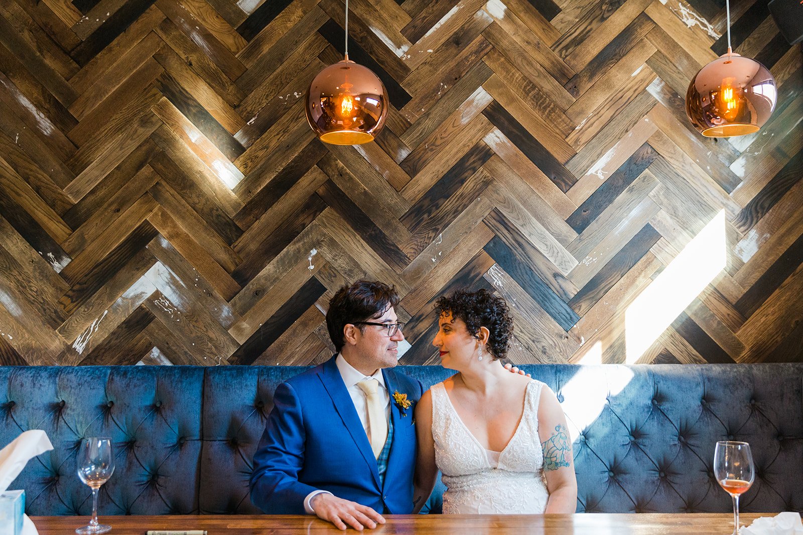  Documentary photo of bride and groom sitting In a booth looking at each other before nontraditional queer, Jewish and Venezuelan wedding ceremony at The Joinery Chicago in Logan Square. 