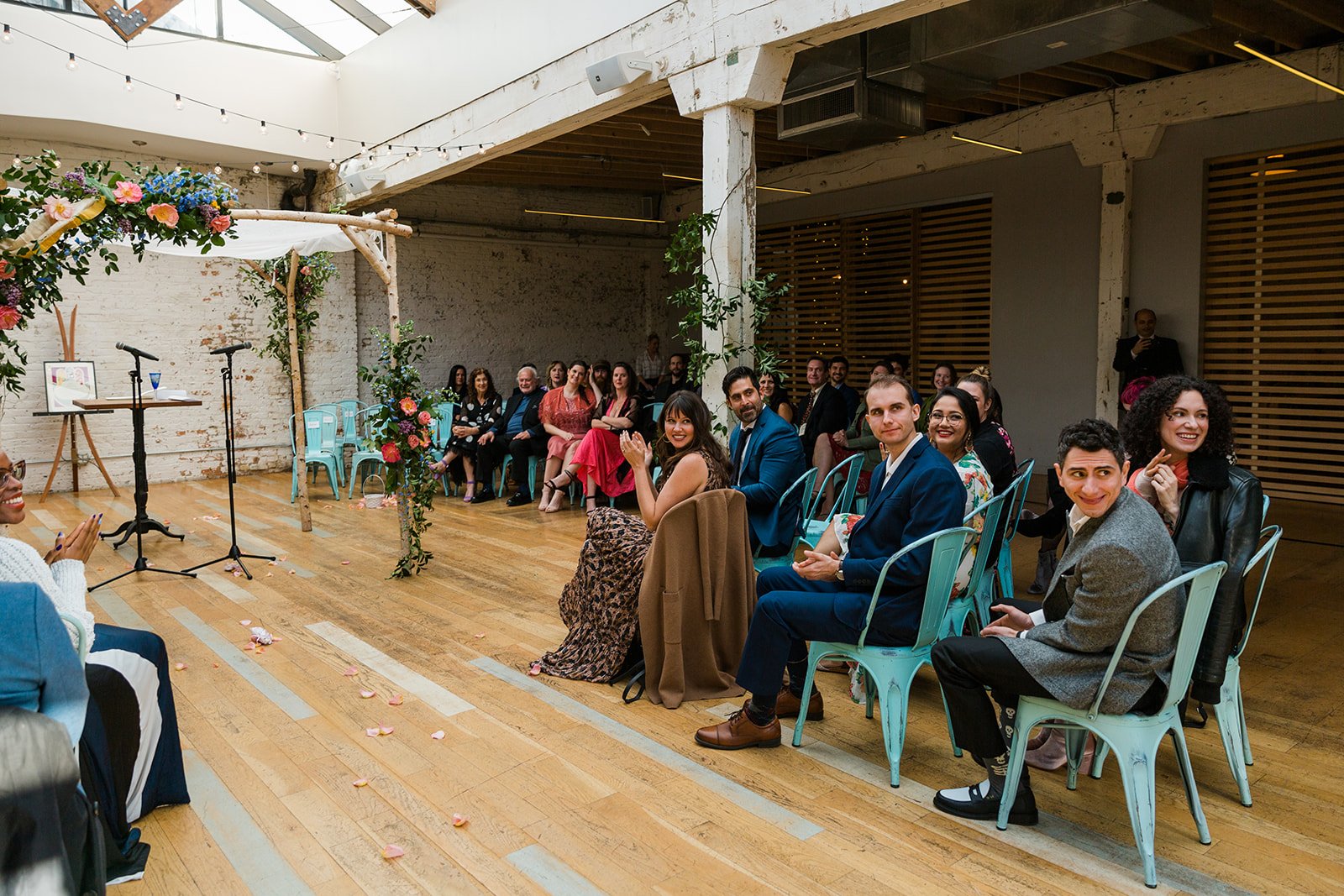  Candid, documentary photo of wedding guests reacting to the couple exiting their nontraditional Jewish and Venezuelan wedding ceremony in the round at The Joinery Chicago an Industrial loft wedding venue In Logan Square. 