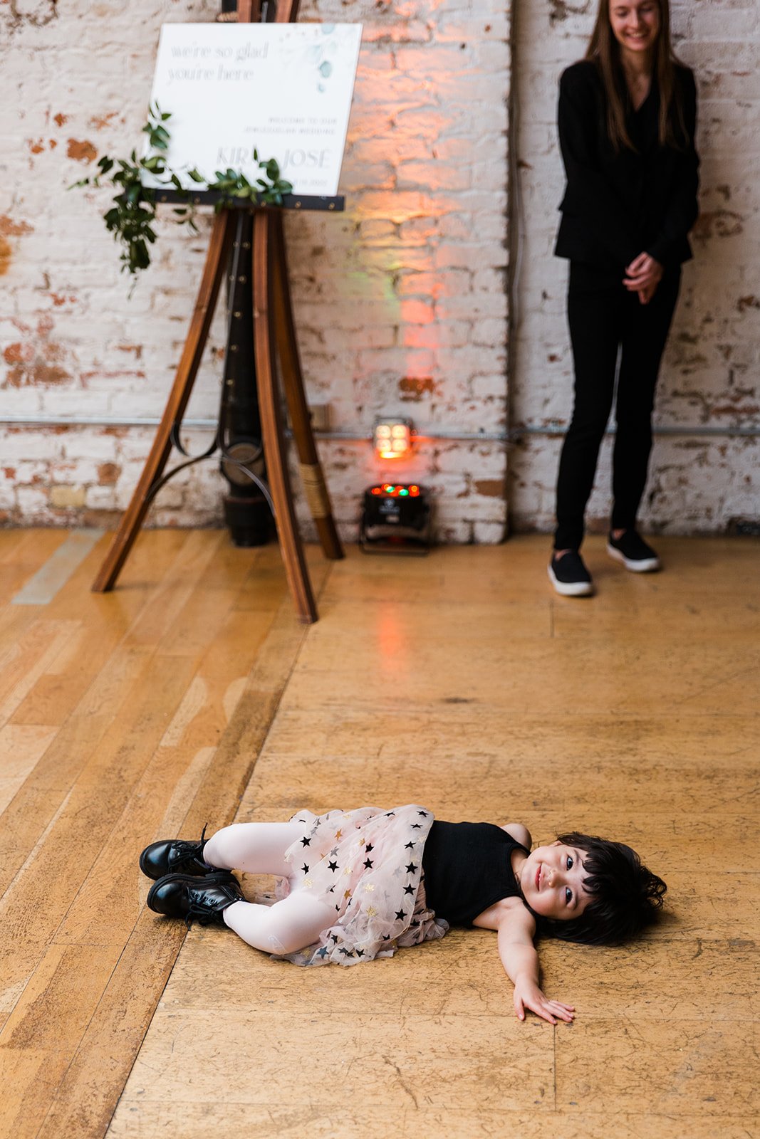  Candid documentary photo of flower girl laying on the floor during cocktail hour after nontraditional Jewish and Venezuelan wedding ceremony at The Joinery Chicago an Industrial loft wedding venue In Logan Square. 