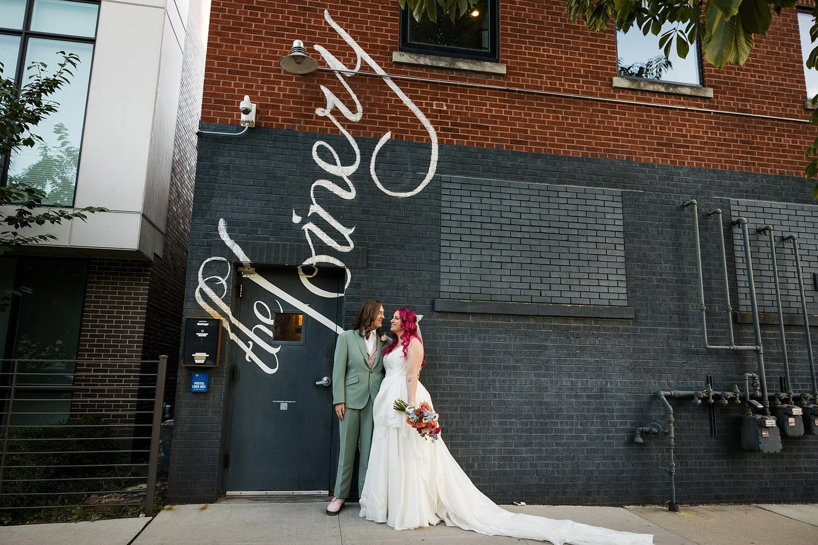  Portrait of nontraditional, queer brides, one in a white dress the other in a light green suit in front of The Joinery in Chicago. A great LGBTQ friendly venue.  