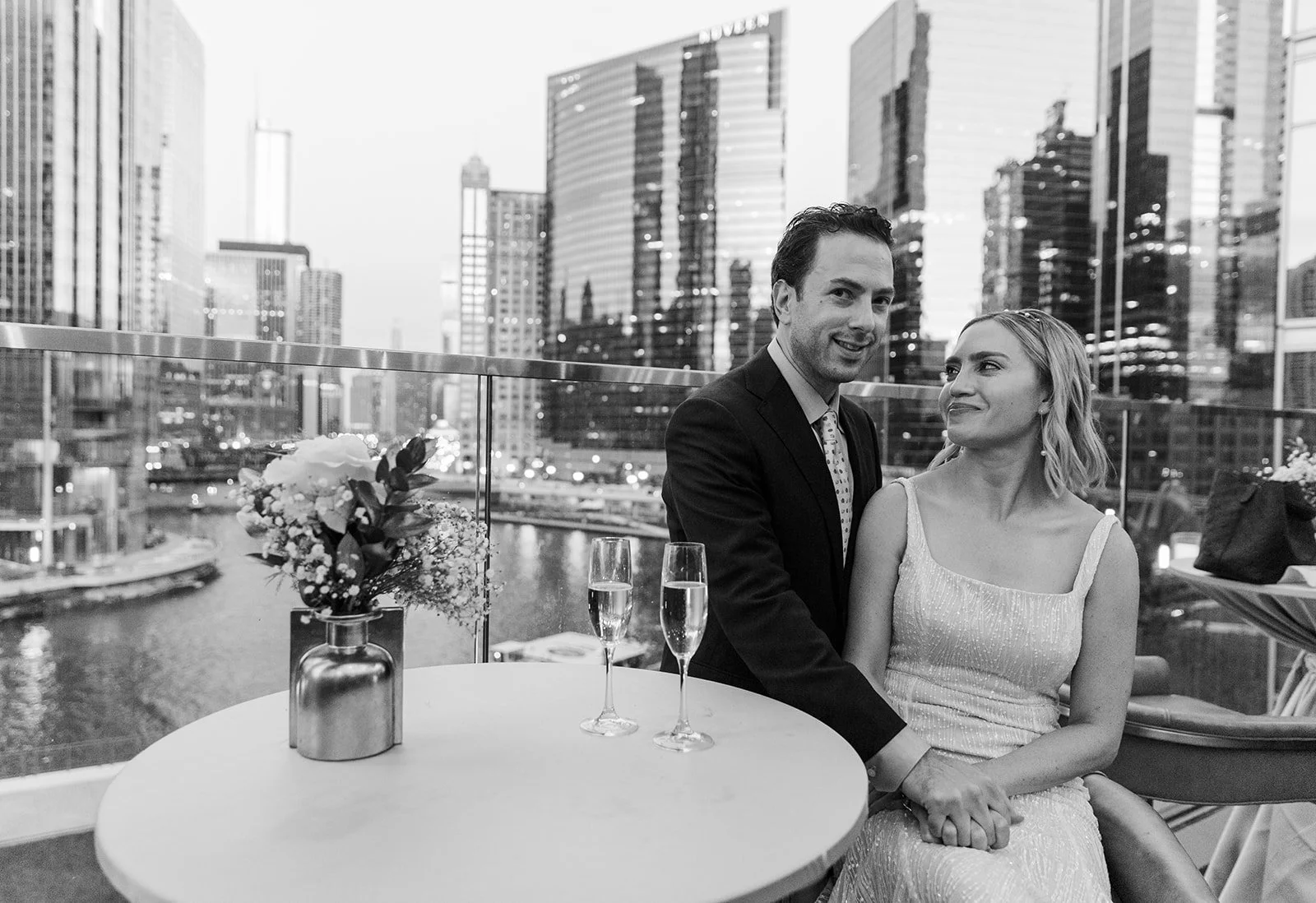  Black and white, documentary style, candid photo of bride and groom on the patio of Gibsons Italia for their nontraditional cocktail style wedding reception. View of the Chicago downtown river skyline behind the. 