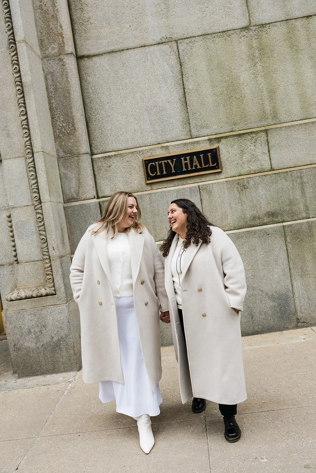 Two brides dressed in white hold hands smiling at each other outside of Chicago's City Hall after their winter elopement. Documentary style wedding photography by queer LGBTQ photographer Fuller Photo Co.