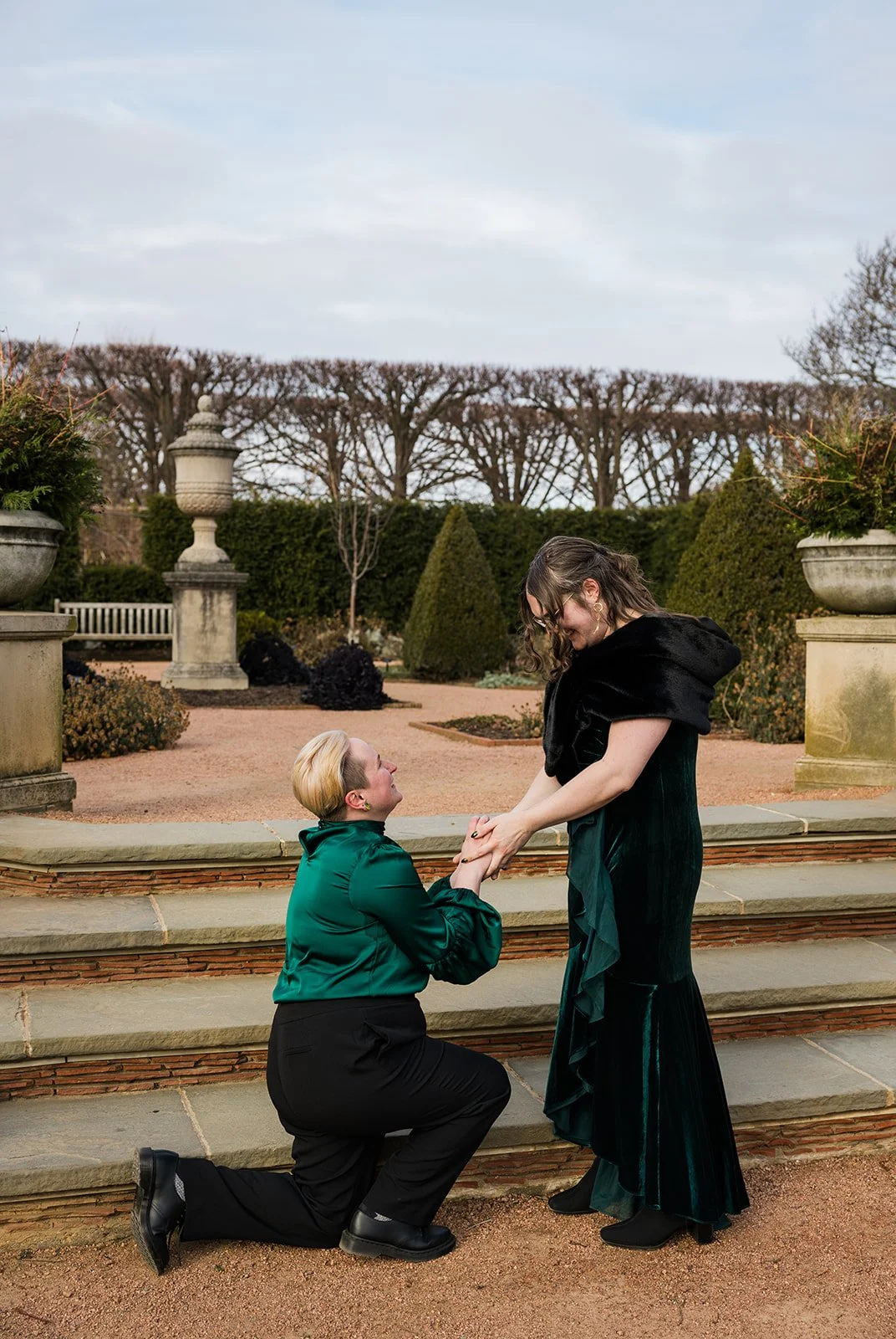 Documentary style photo of a queer couples surprise proposal on a sunny winter day at The Chicago Botanical Garden. One partner is down on their knees as the other holds their hand.