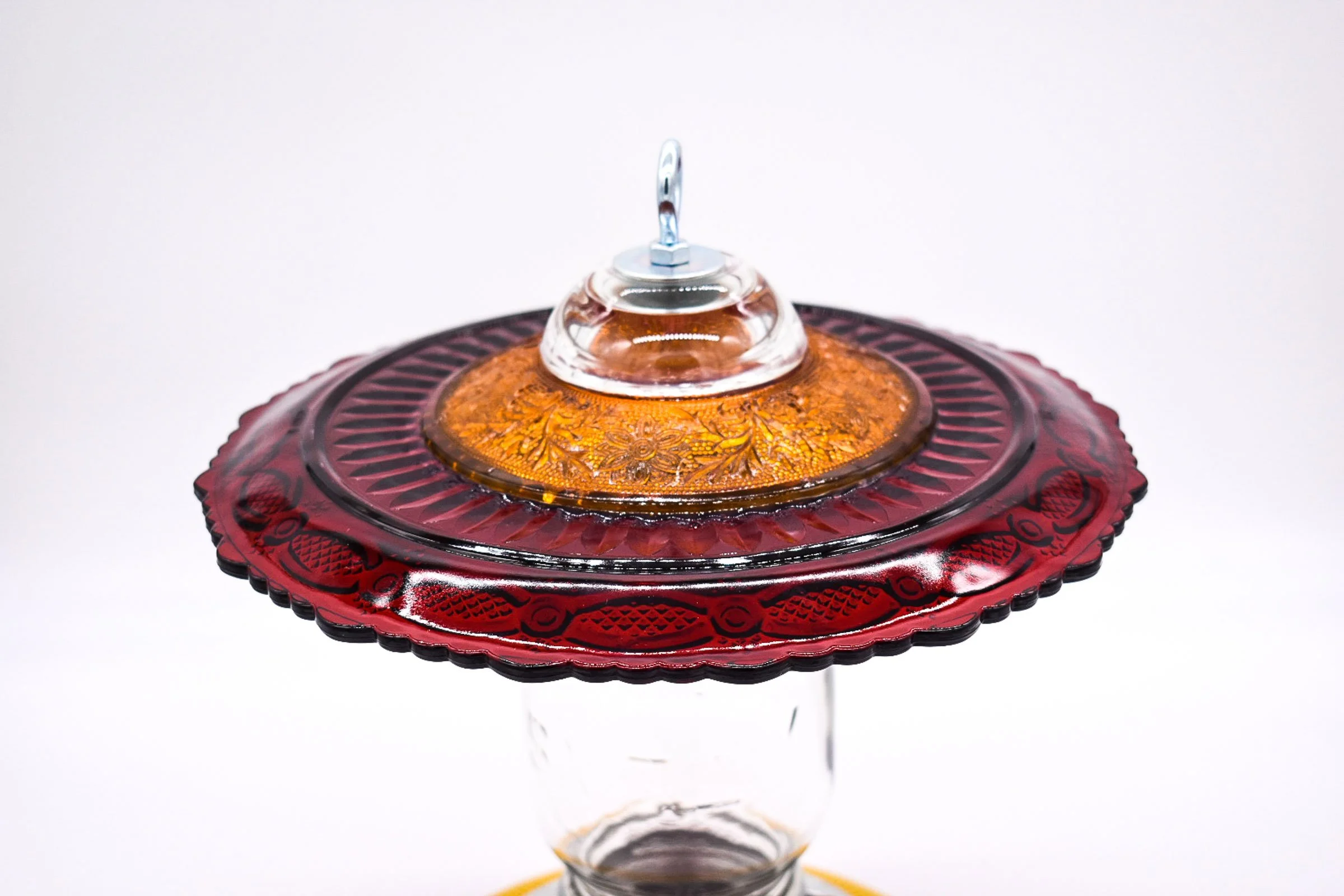 Angled close-up of the handmade bird feeder showing the raised ruby red glass pattern, amber glass saucer, and silver hanging attachment.
