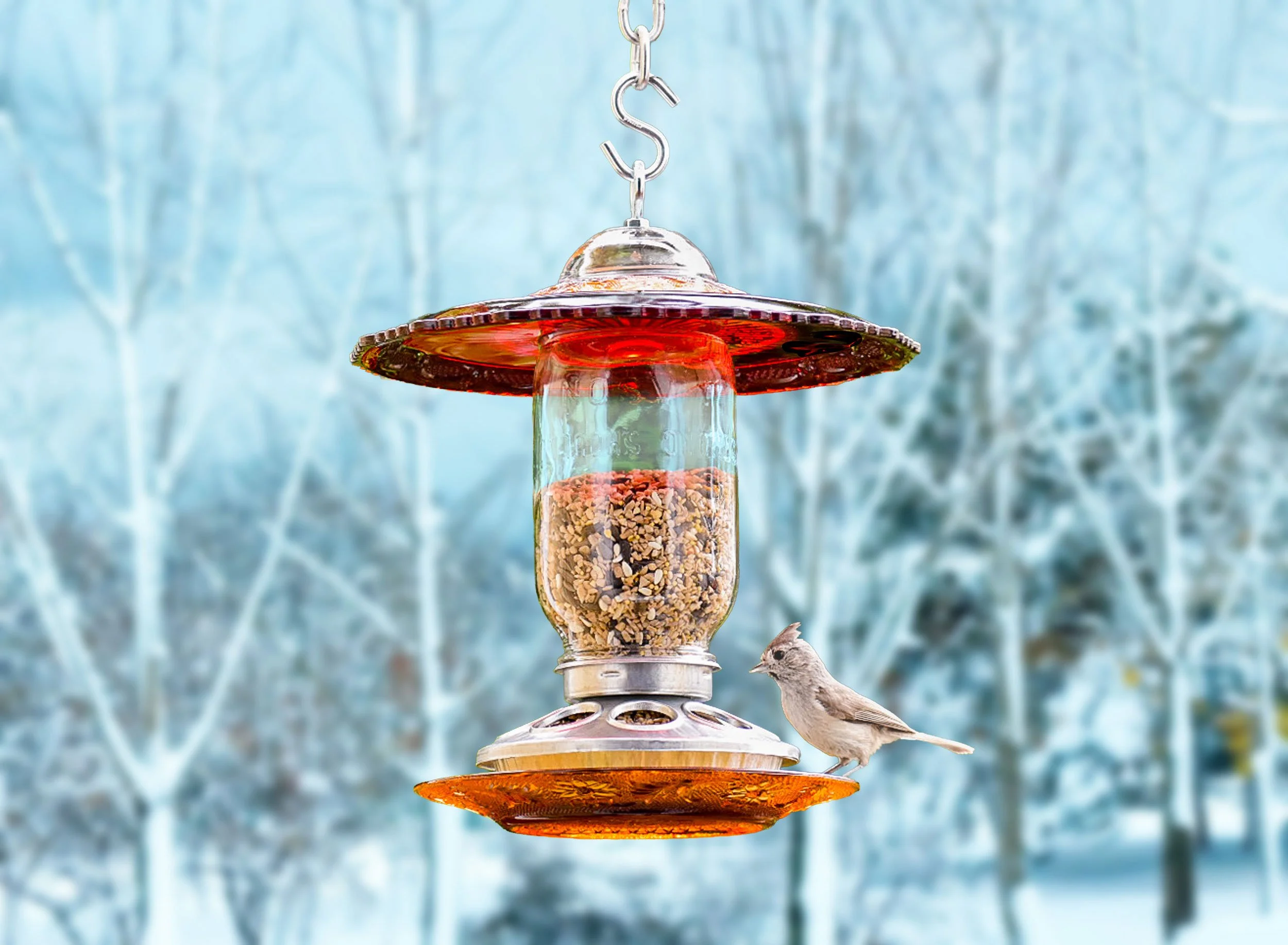 Ruby red and amber glass seed bird feeder hanging in a snowy winter setting with a small bird perched beside the galvanized metal feeding base.
